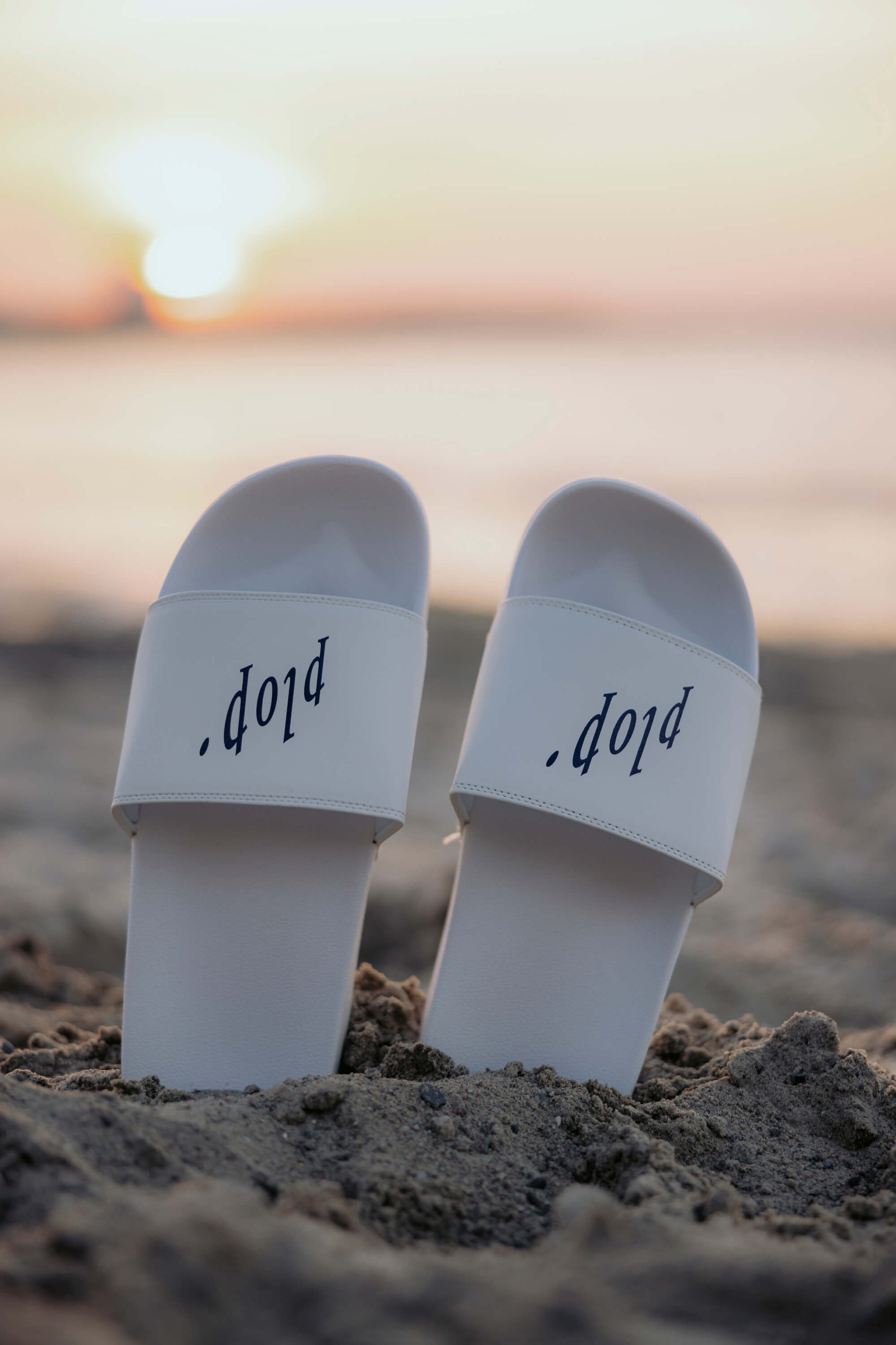 Weisse Fördeslipper mit dem Schriftzug "plop." stecken hochkant im Sand. Der Sonnenuntergang ist im Hintergrund unscharf zu sehen.