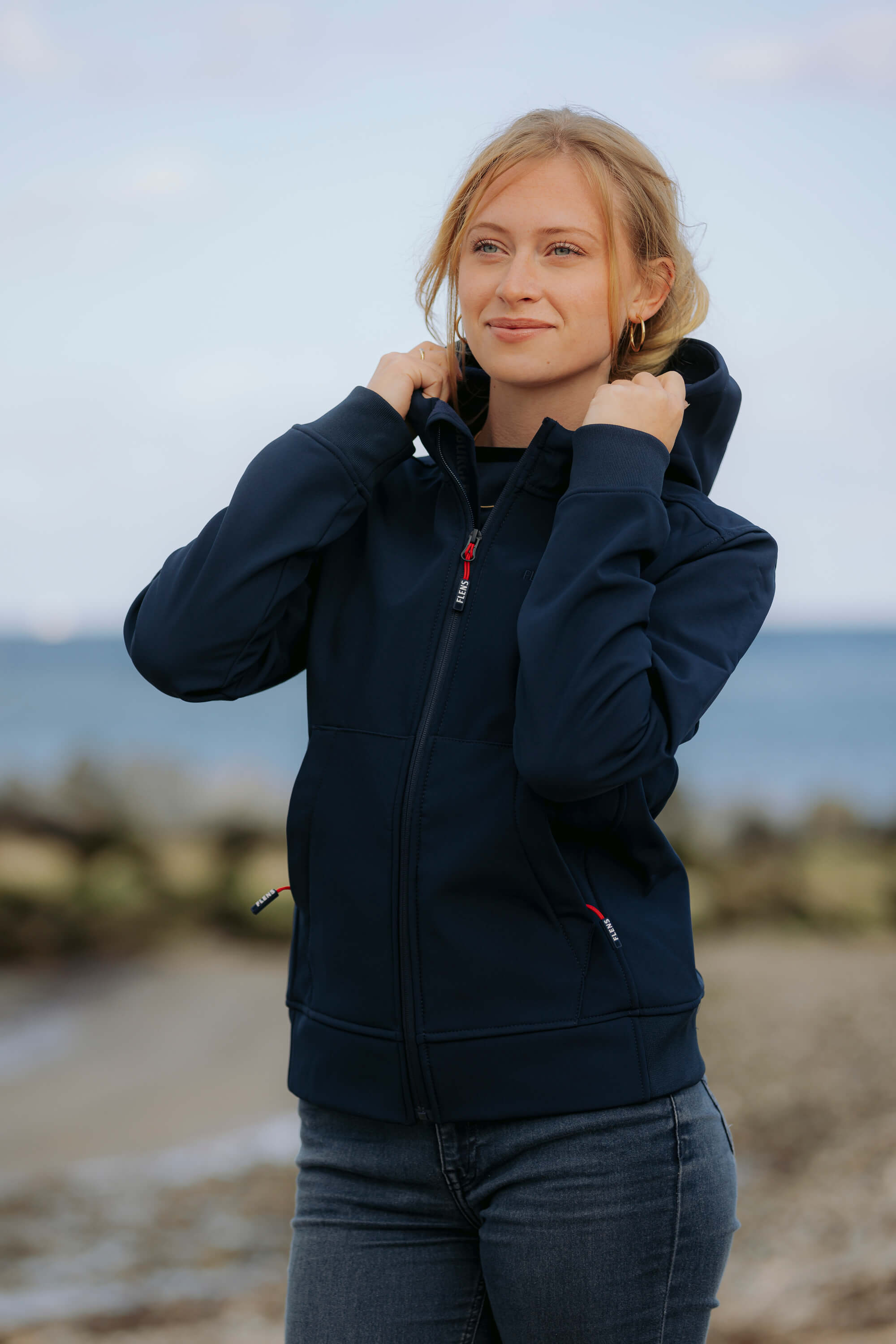 Eine junge Frau trägt die dunkelblaue FLENS Softshelljacke und hält die Kapuze mit beiden Händen fest, während sie am Strand steht.