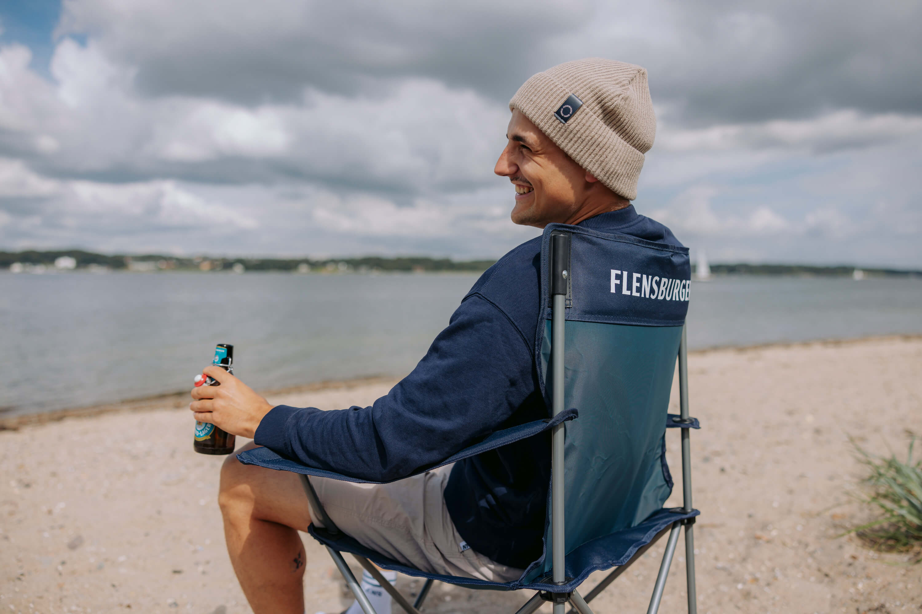 Junger Mann sitzt in einem Campingstuhl am Strand. Er hält eine Flasche in der Hand und blickt lächelnd auf das Wasser. Der Himmel ist bewölkt.