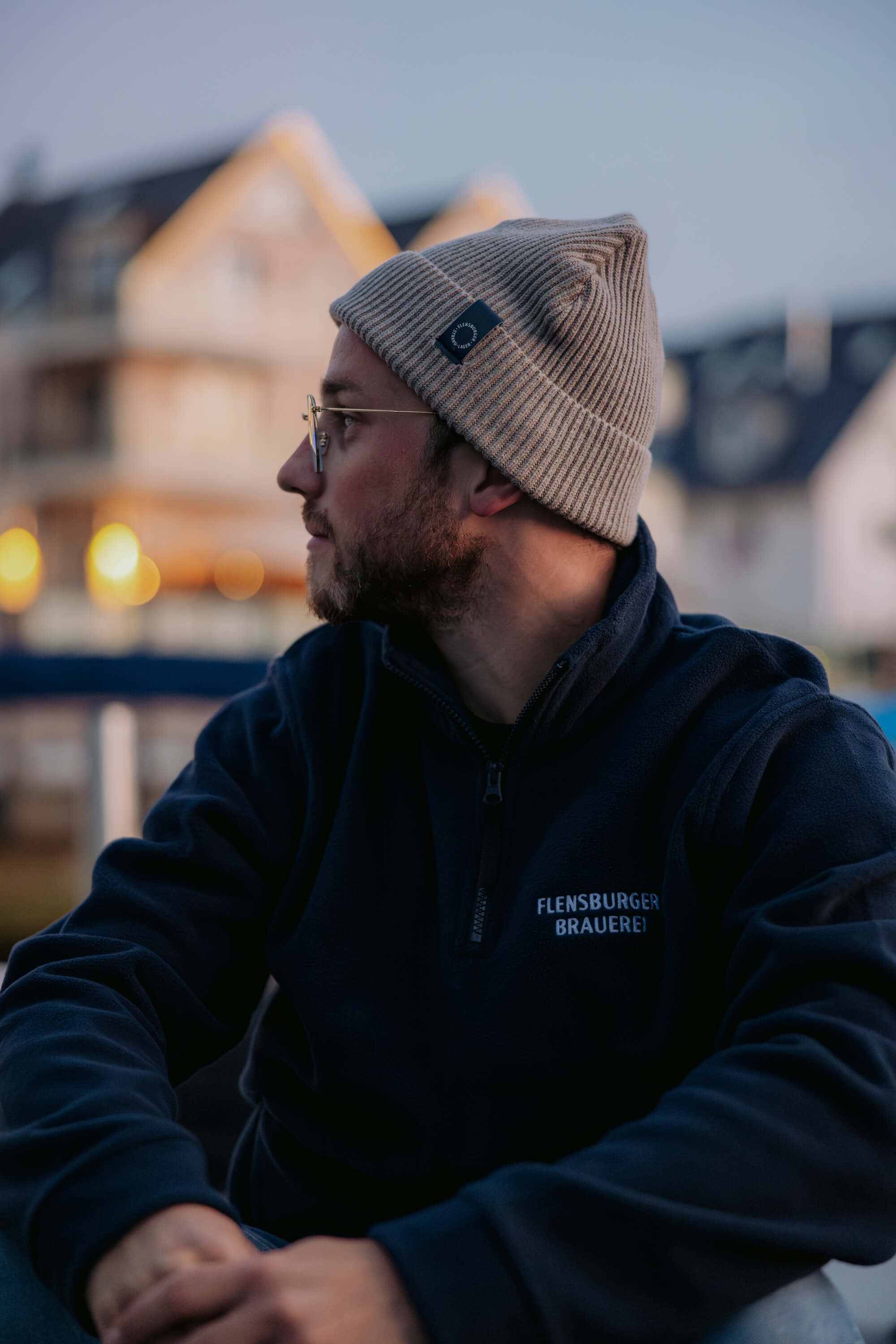 Mann trägt eine beige gestrickte Fisherman Beanie und eine dunkelblaue Jacke mit dem Schriftzug „Flensburger Brauerei“. Er sitzt seitlich und schaut nach links, im Hintergrund sind unscharfe Häuser zu sehen.