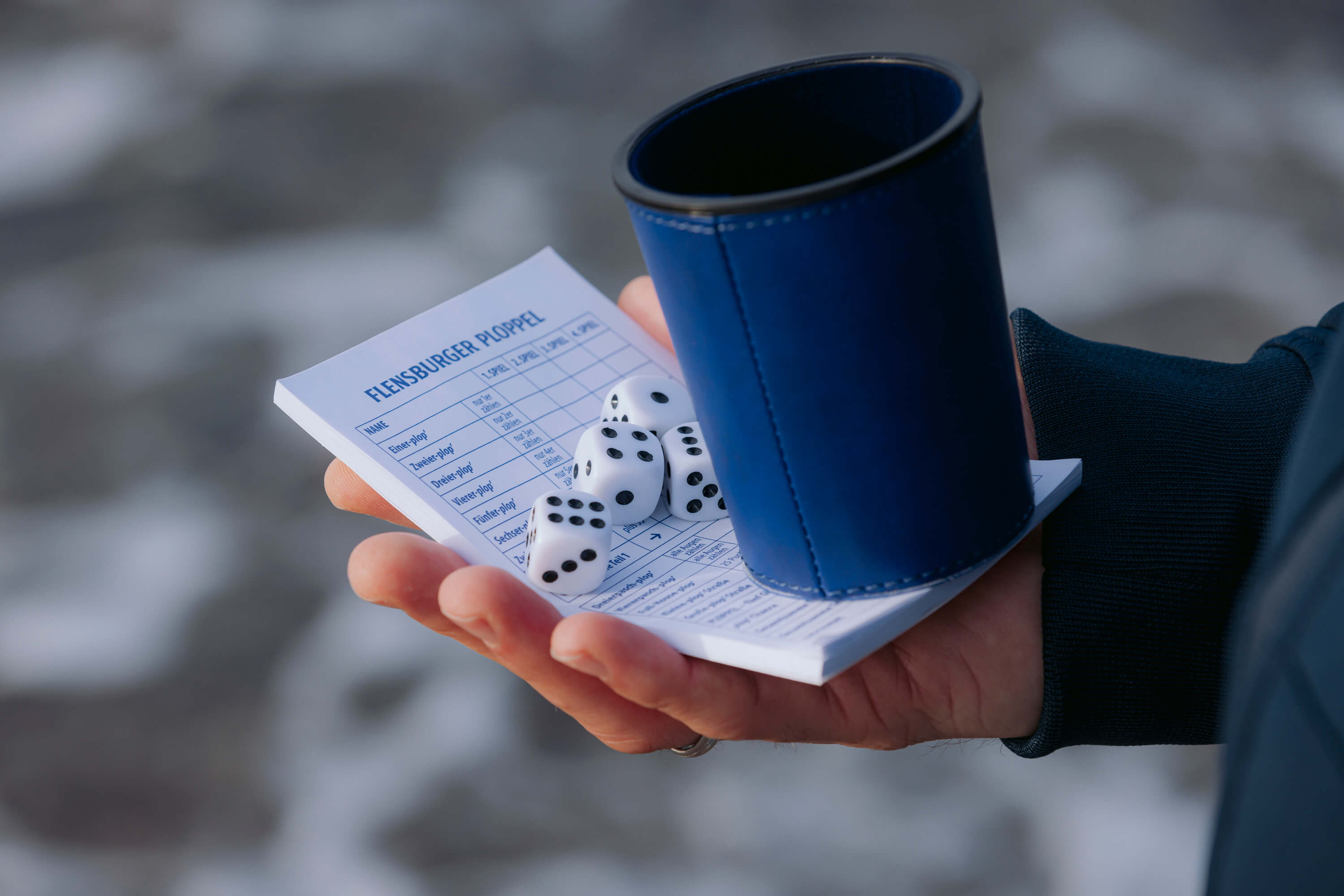Eine Hand hält ein Flensburger Ploppel Würfelset mit fünf weißen Würfeln, einem blauen Würfelbecher und einem Spielzettel mit der Überschrift „FLENSBURGER PLOPPEL“.