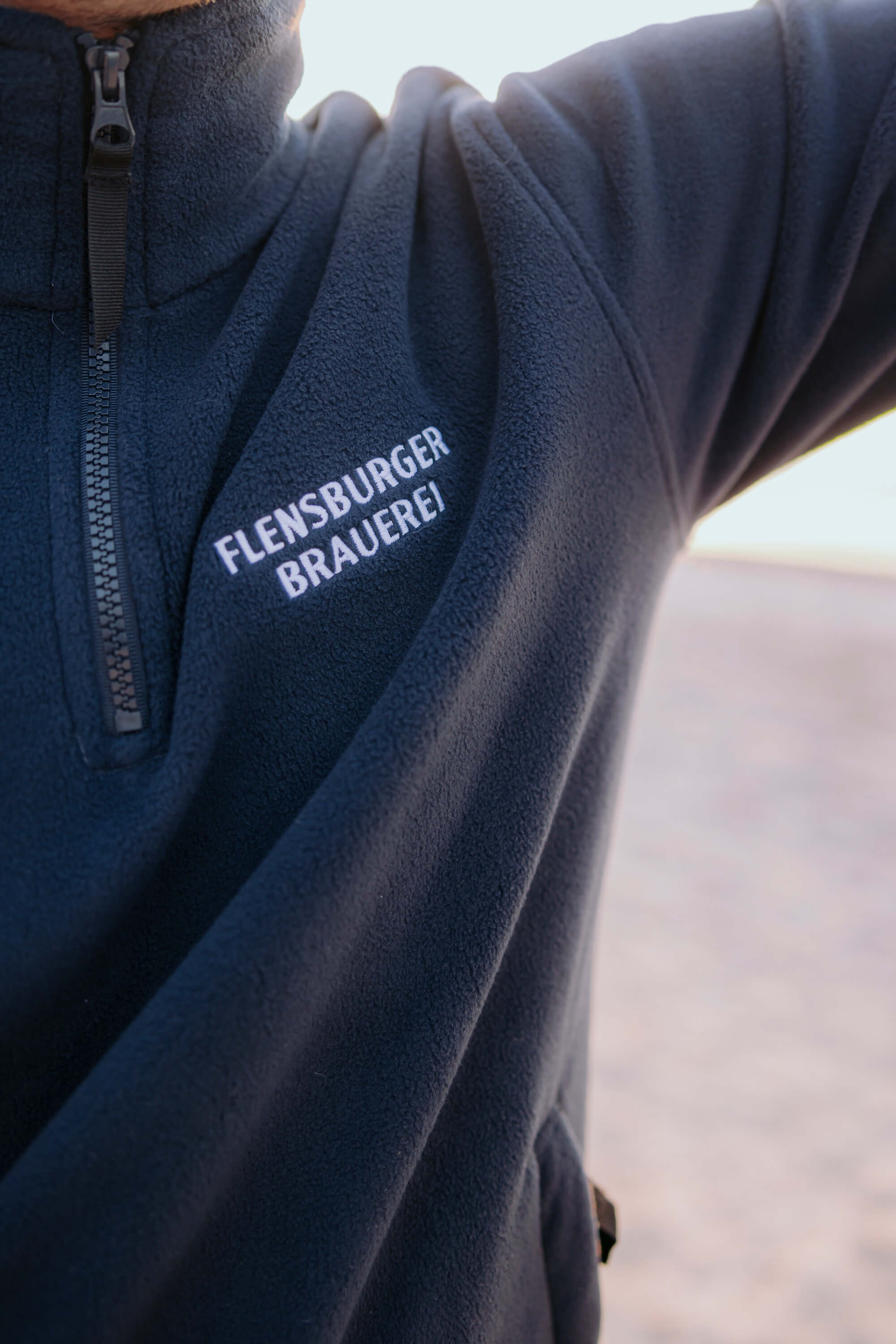 Detailansicht: dunkelblauer Fleece-Pullover mit Reißverschluss und weißem Schriftzug „FLENSBURGER BRAUEREI“ auf der Brust.