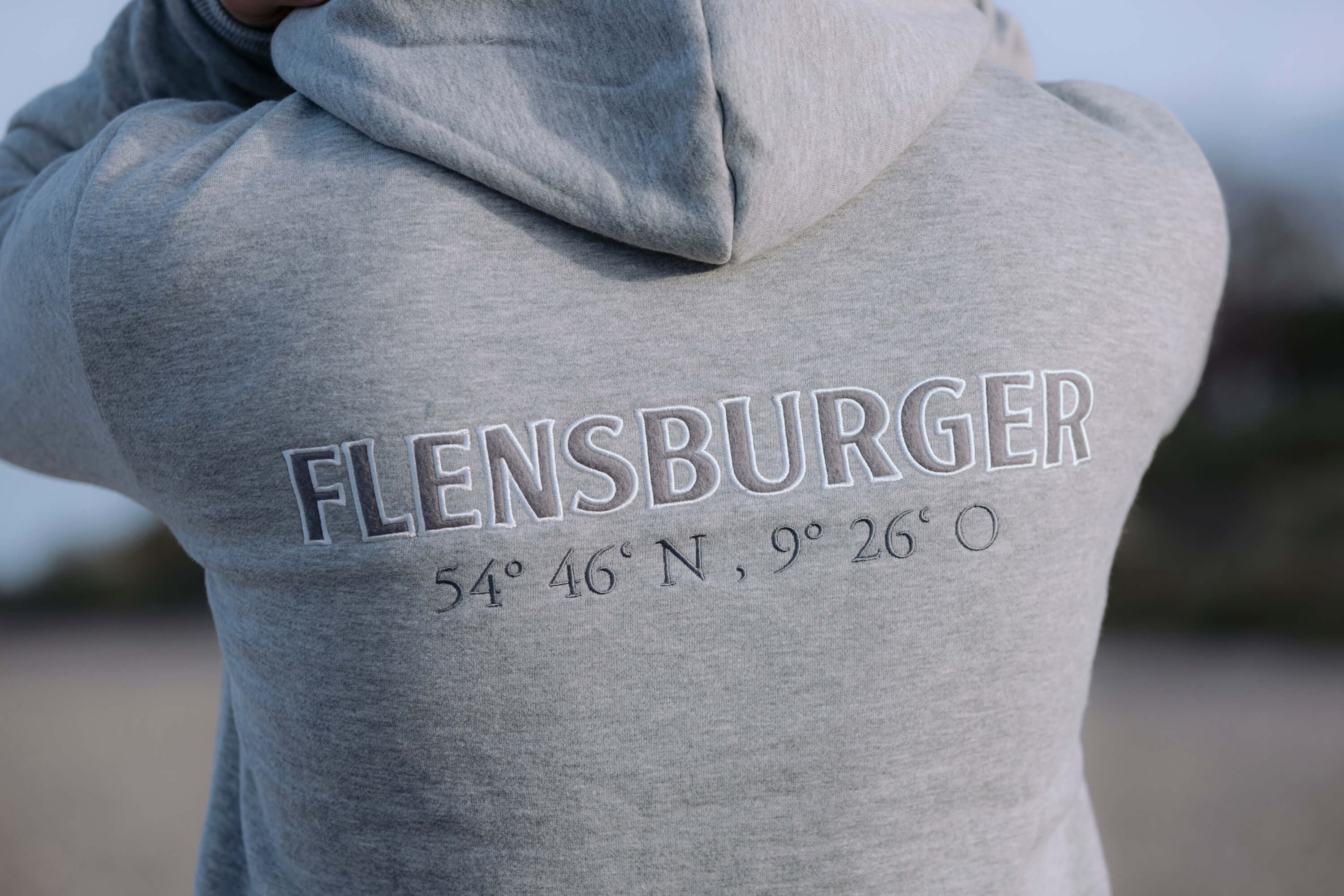 Hellgraue Sweatshirtjacke mit Kapuze. Auf dem Rücken steht in großen weißen Buchstaben "FLENSBURGER". Darunter sind die Koordinaten "54° 46‘ N, 9° 26‘ O" in schwarzer Schrift aufgestickt.