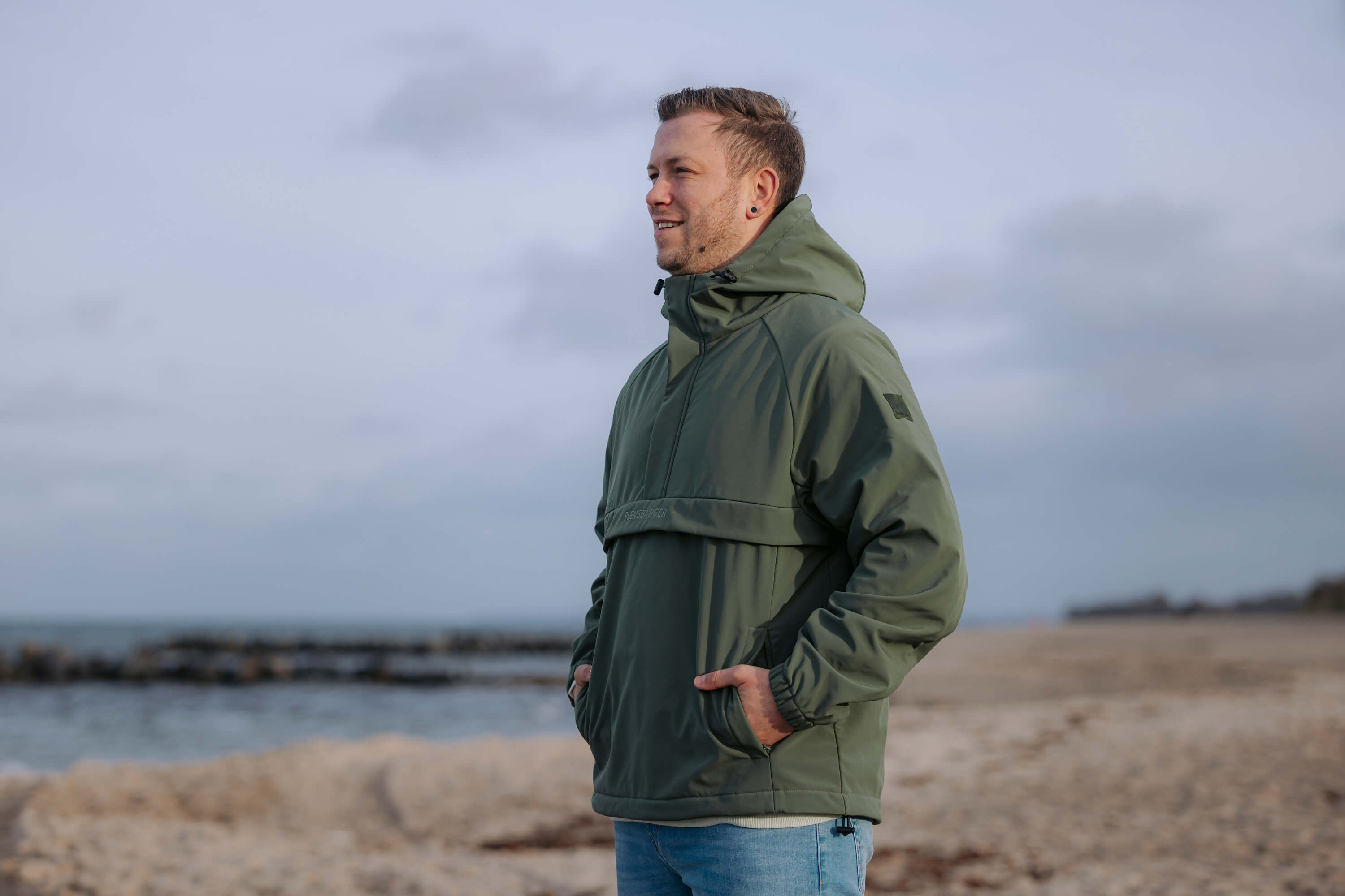 Mann trägt den wetterfesten Windbreaker "Skagerrak" in Olivgrün und steht mit den Händen in den Taschen am Strand neben dem Meer.