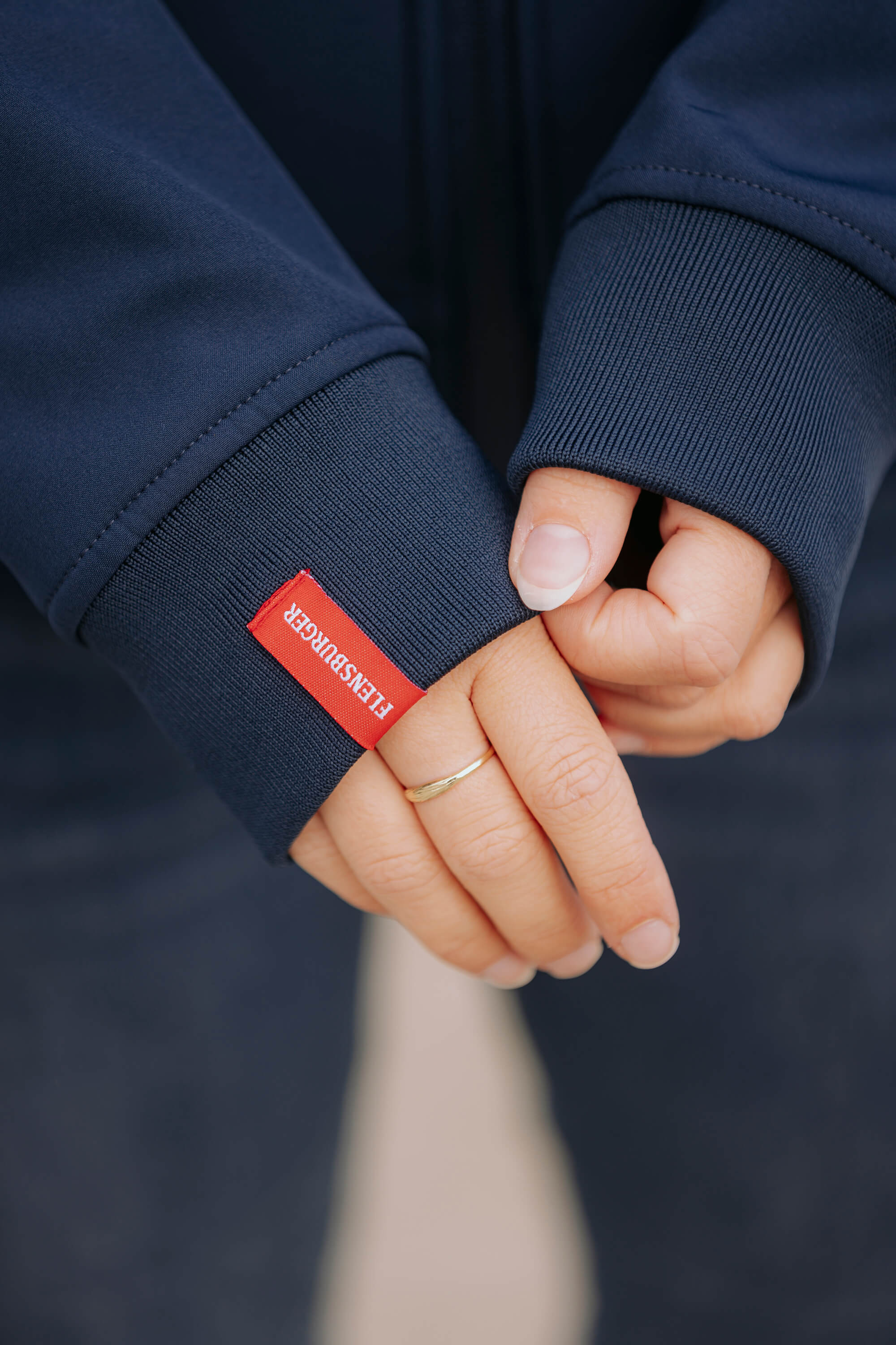 Nahaufnahme von Händen in dunkelblauer Softshelljacke mit rotem Label "FLENSBURGER" am Ärmel. Eine Hand hält den Daumen der anderen.