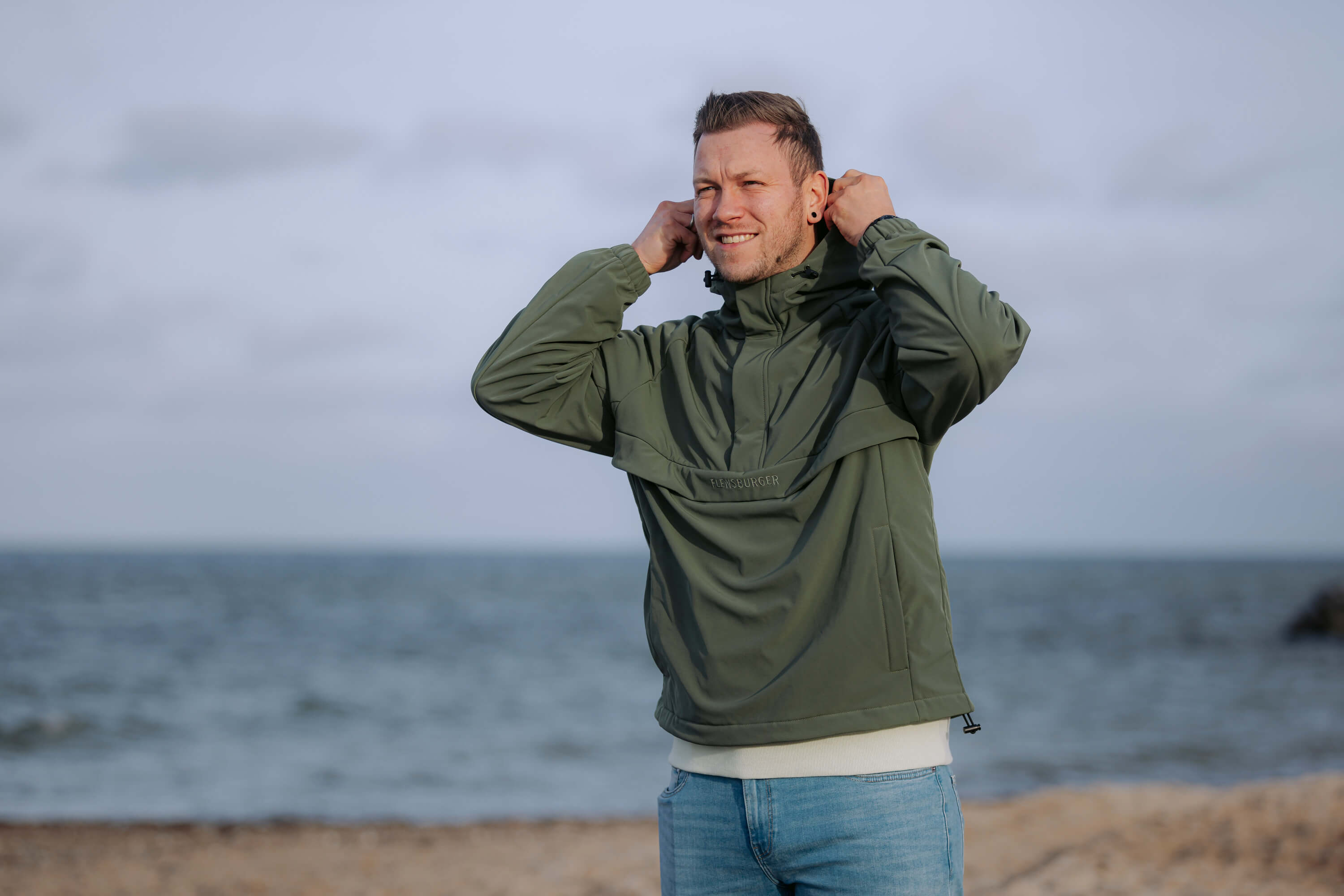 Ein Mann zieht die Kapuze seines wetterfesten, olivgrünen Windbreakers mit der Aufschrift „FLENSBURGER“ am Strand auf.