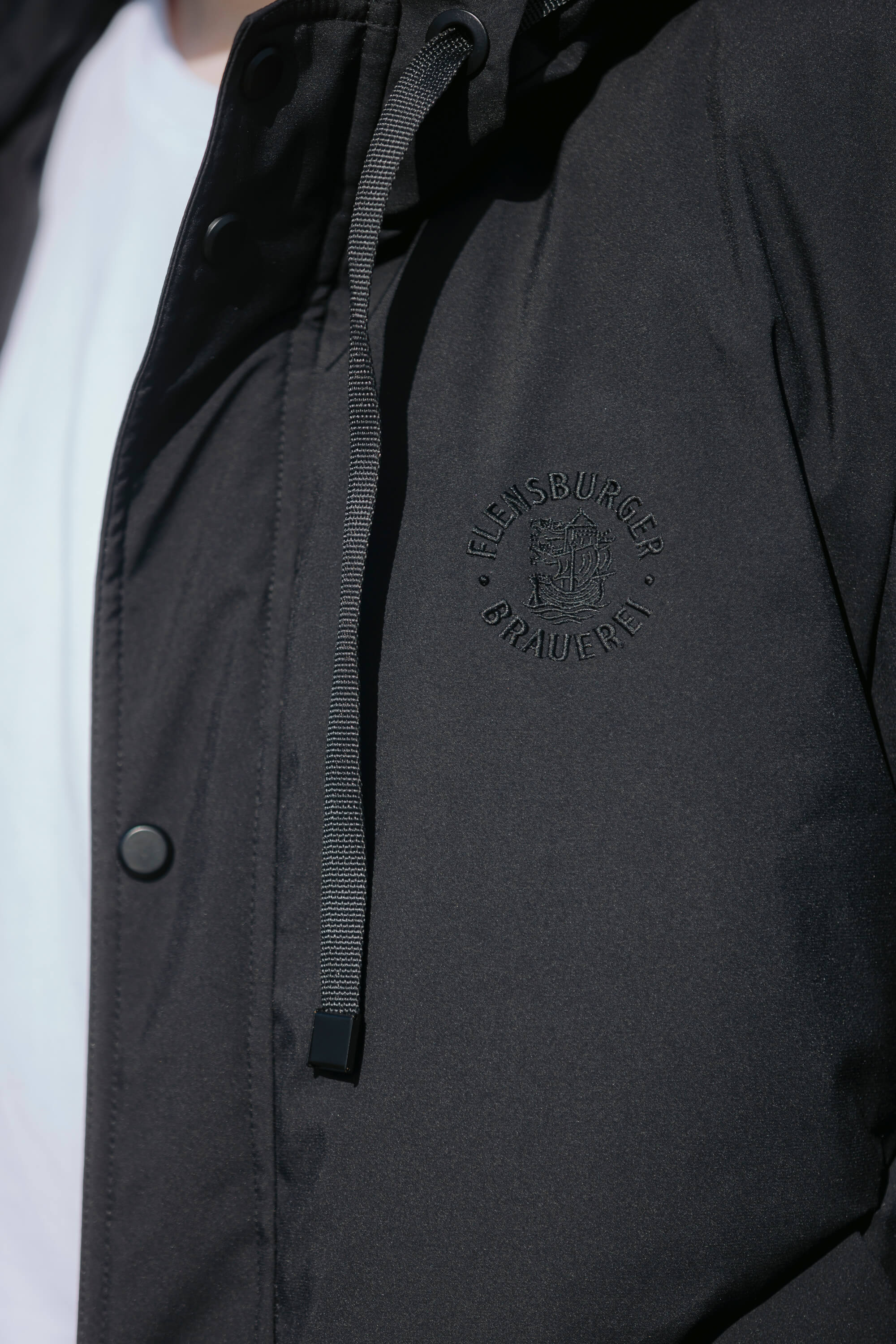 Detailansicht: Schwarzer Winterparka mit Kapuze zeigt einen schwarzen Kordelzug und eine auf der Brust gestickte, schwarze Schrift “Flensburger Brauerei” mit einem Wappen.