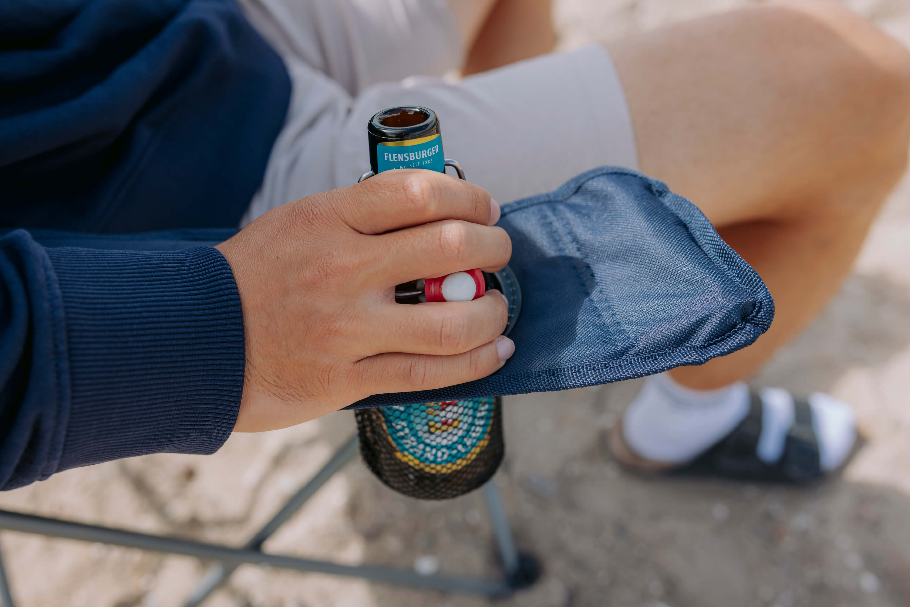 Eine Hand hält eine Flasche Flensburger Bier auf der Armlehne eines Campingstuhls. Der Stuhl hat eine blaue, gewebte Hülle und steht auf sandigem Untergrund.