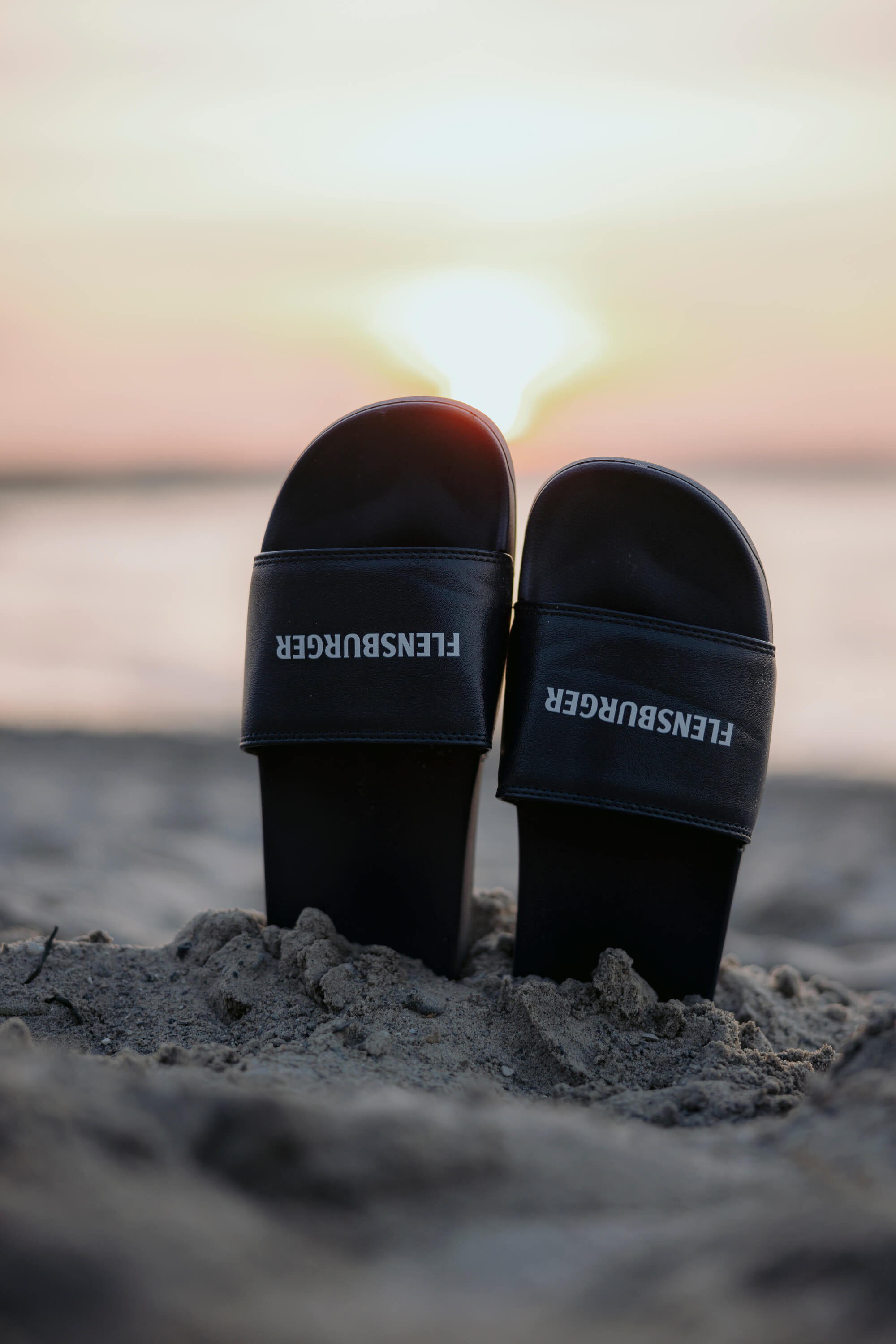 Schwarze Fördeslipper "Flensburger" stecken in aufrechter Position im Sand. Der Text "FLENSBURGER" ist weiß und auf beiden Slippern deutlich lesbar. Im Hintergrund ist ein Sonnenuntergang am Meer zu sehen.