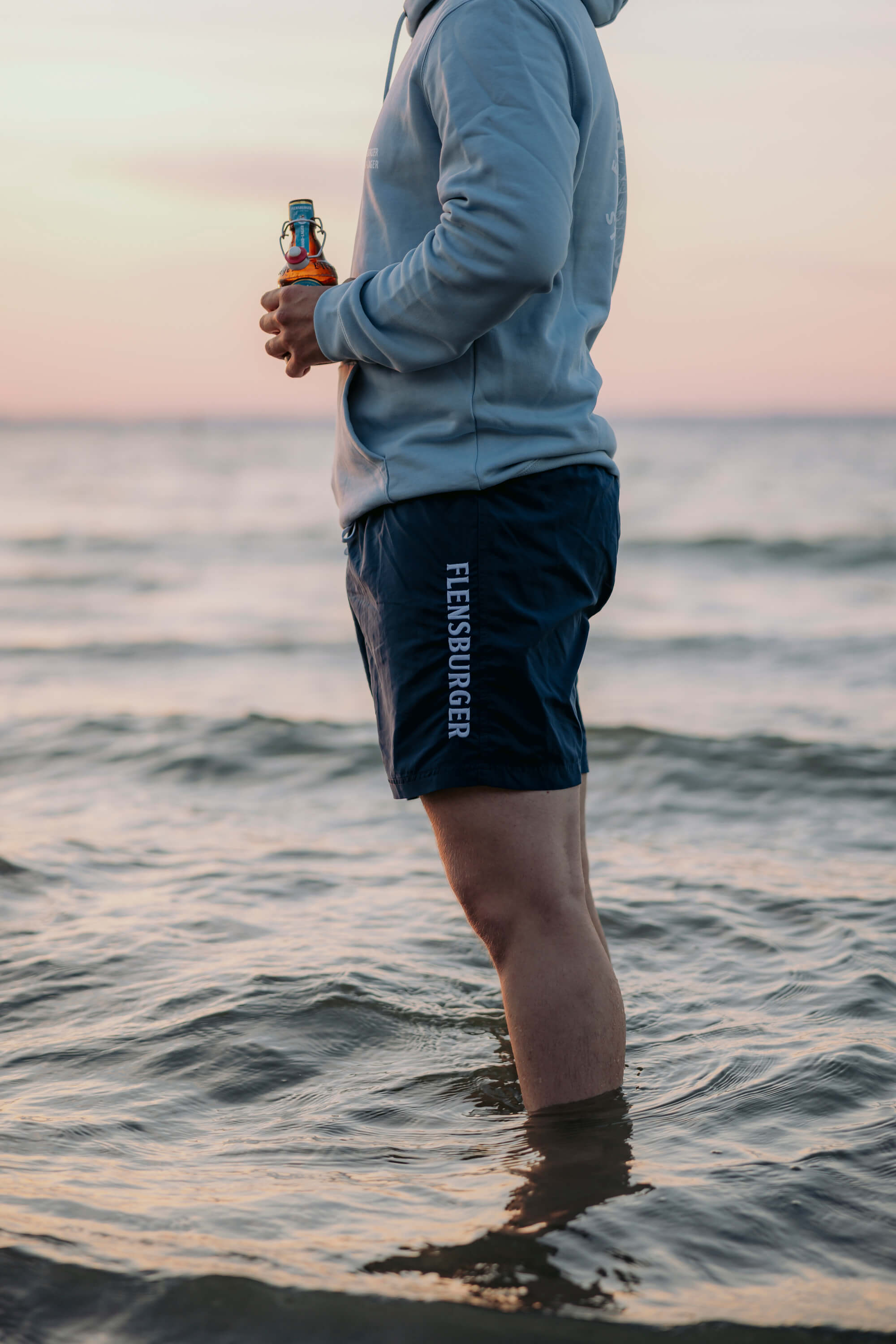 Eine Person steht mit den Beinen im Wasser. Die Person trägt eine hellblaue Kapuzenjacke und dunkelblaue Badehose mit weißem "FLENSBURGER"-Schriftzug. Die Person hält eine Flasche in beiden Händen.