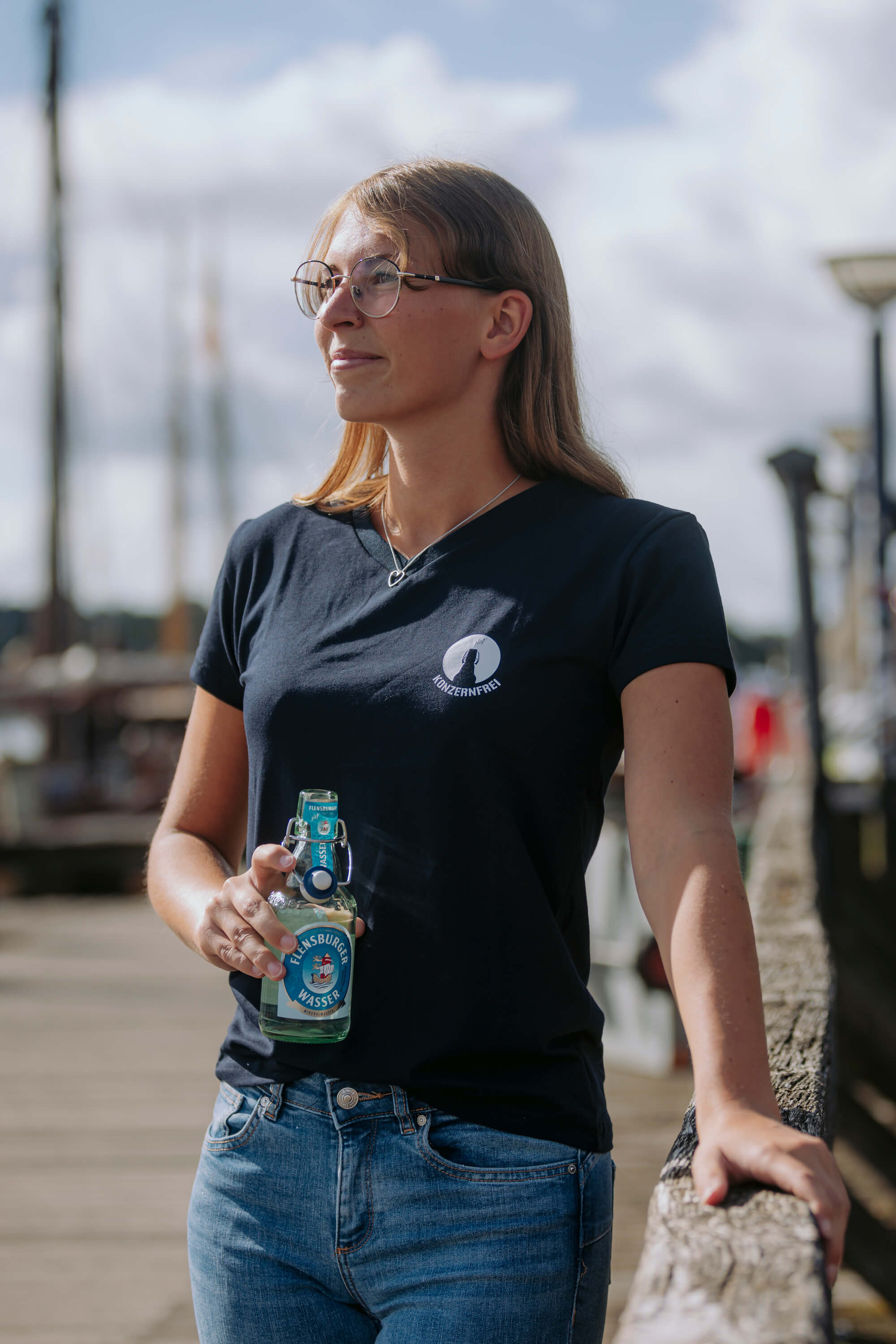 Frau trägt ein navyfarbenes Damen-Shirt mit V-Ausschnitt und weißem „konzernfrei“-Logo auf Herzhöhe. Sie hält eine Flasche „Flensburger Wasser“ in der Hand und steht an einem Steg.