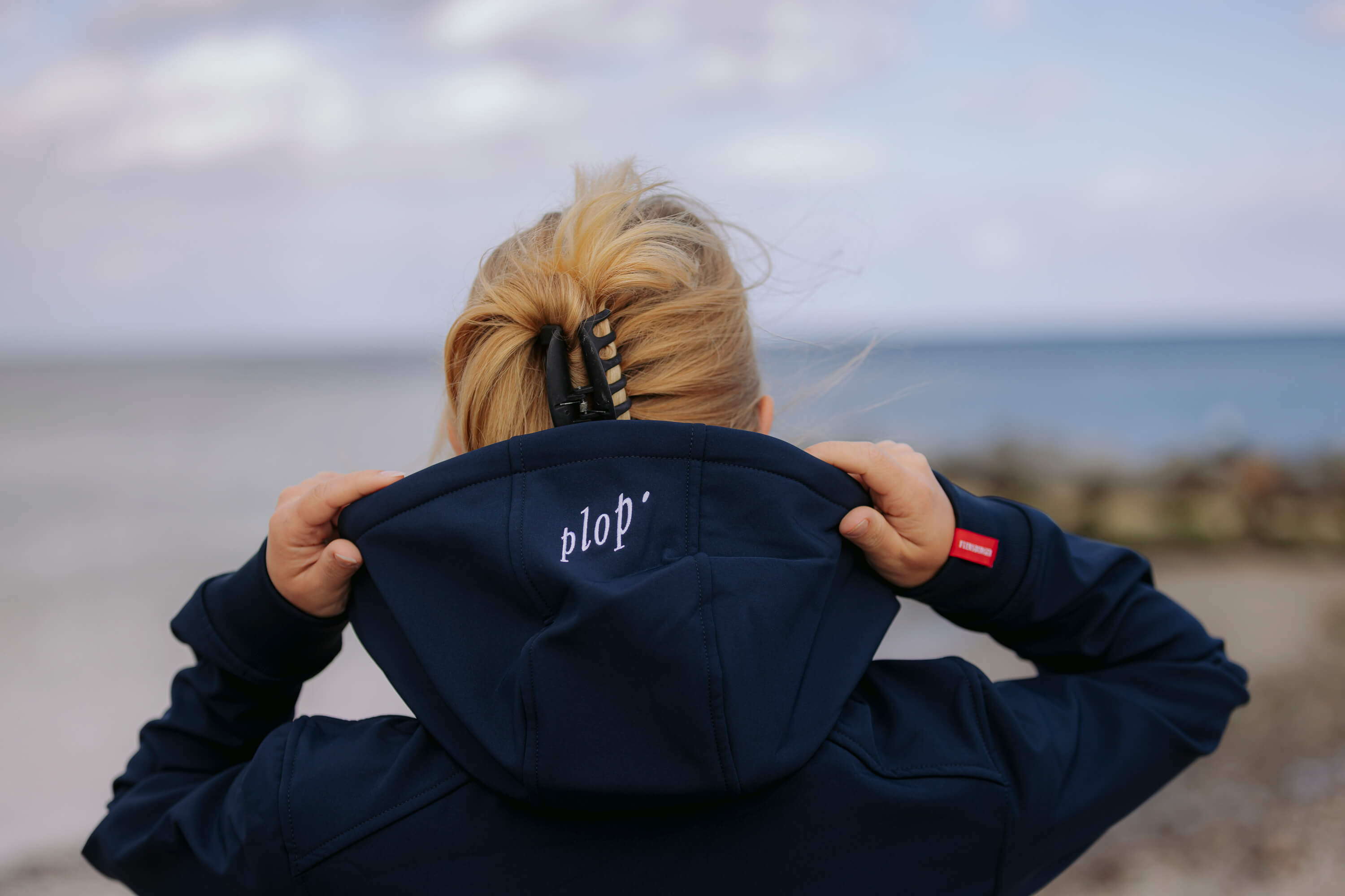 Eine Frau zieht die Kapuze einer navyblauen Softshelljacke hoch. Auf der Kapuze steht der weiße Schriftzug „plop’“. Die Frau trägt eine schwarze Haarspange, das Meer ist im Hintergrund sichtbar.