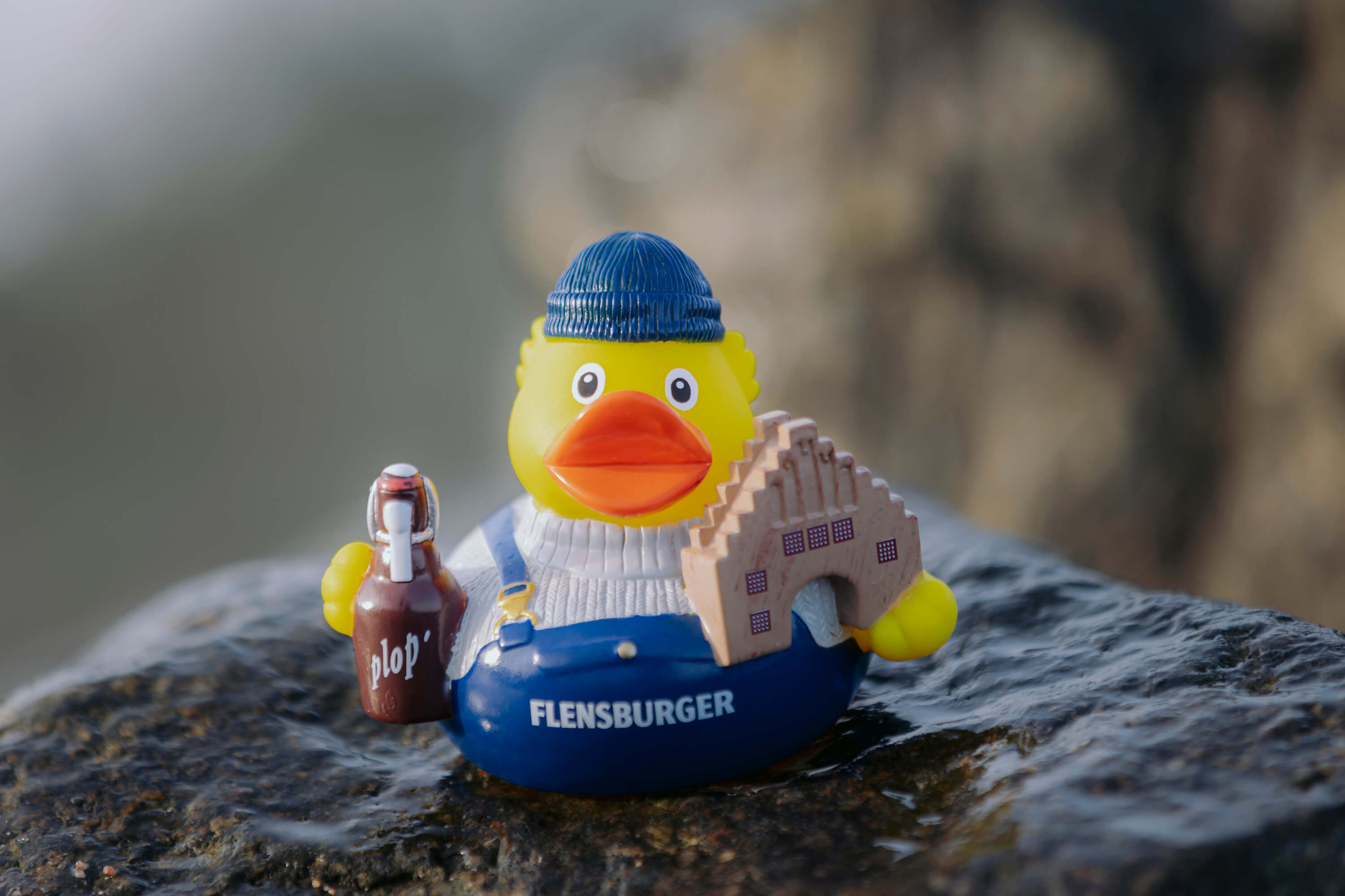 Eine gelbe Gummiente mit blauer Mütze und blauer Latzhose mit dem Schriftzug „FLENSBURGER“. Sie hält eine Brauerei-Flasche mit der Aufschrift „plop“ und ein Modell des Nordertors.