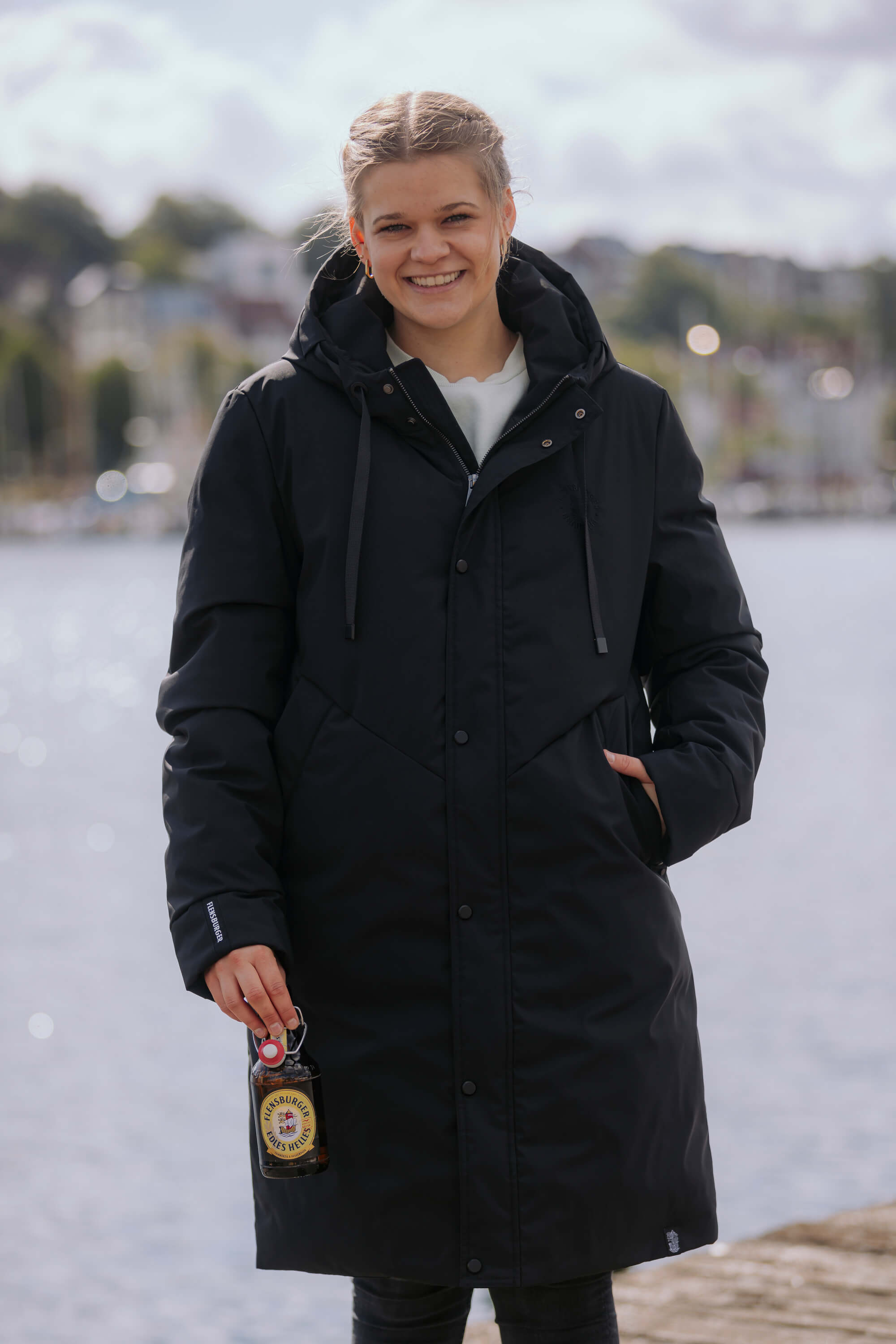Frau trägt den schwarzen Winterparka FLENS "Ruben" und hält eine Flasche Flensburger Edles Helles in der rechten Hand. Sie steht vor einem Gewässer und lächelt in die Kamera.