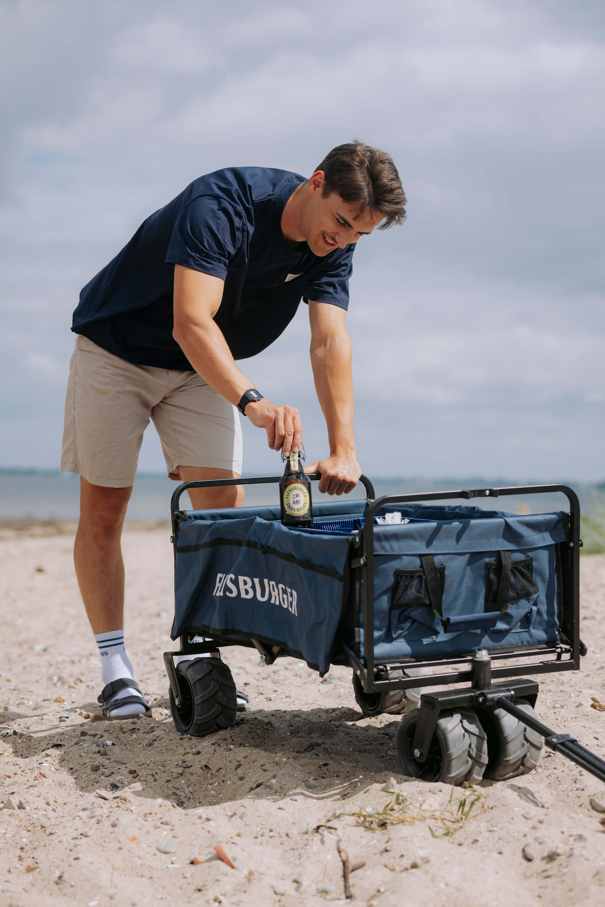 Mann in Shorts und T-Shirt stellt eine Flasche Flensburger Pilsener in einen blauen faltbaren Bollerwagen mit breiten Reifen und Aufschrift „FLENSBURGER“.