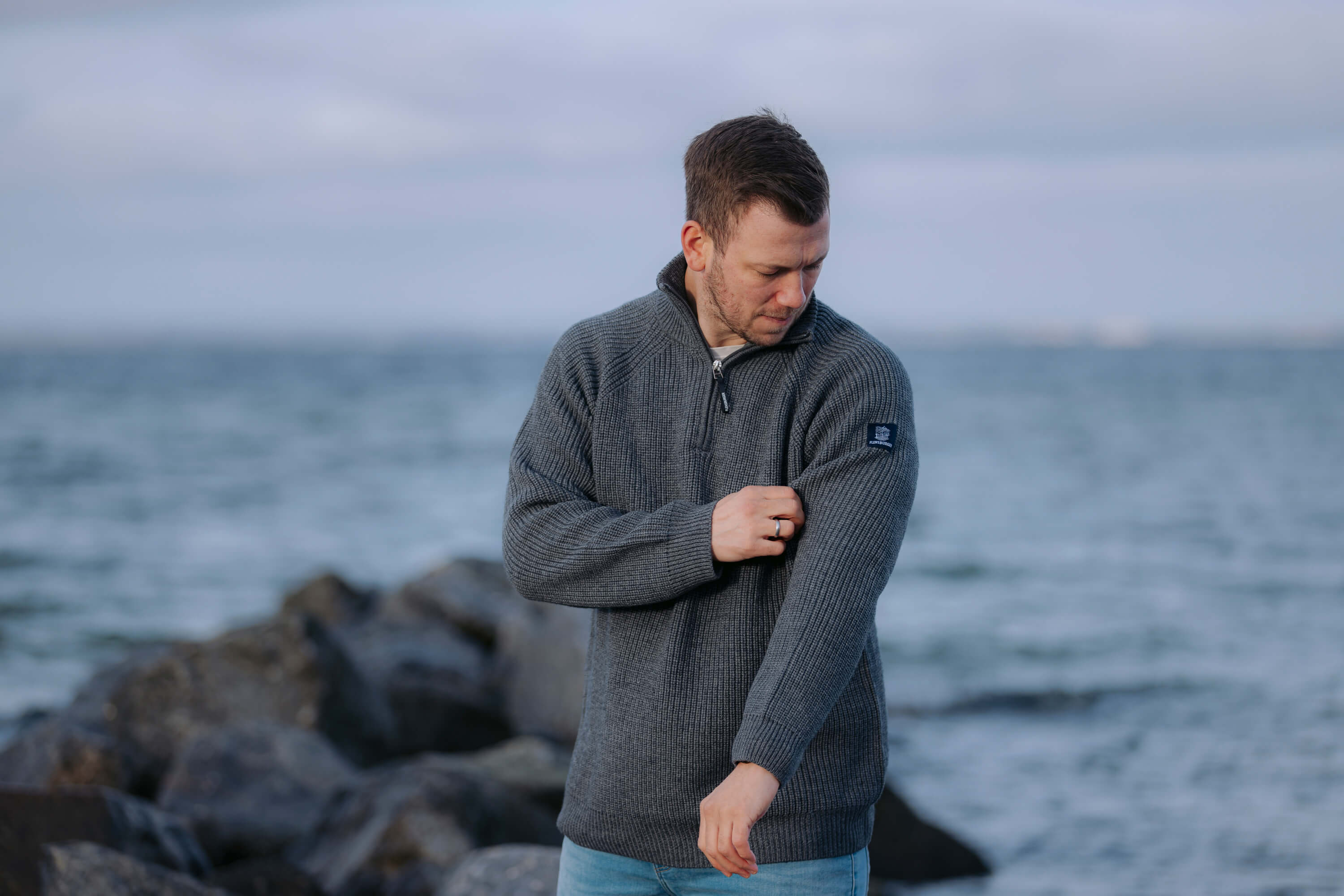 Mann trägt anthrazitfarbenen, halbhoch gezippten Strickpullover mit Logo auf dem linken Oberarm und krempelt den rechten Ärmel hoch. Er steht vor felsiger Küste und Meer im Hintergrund.