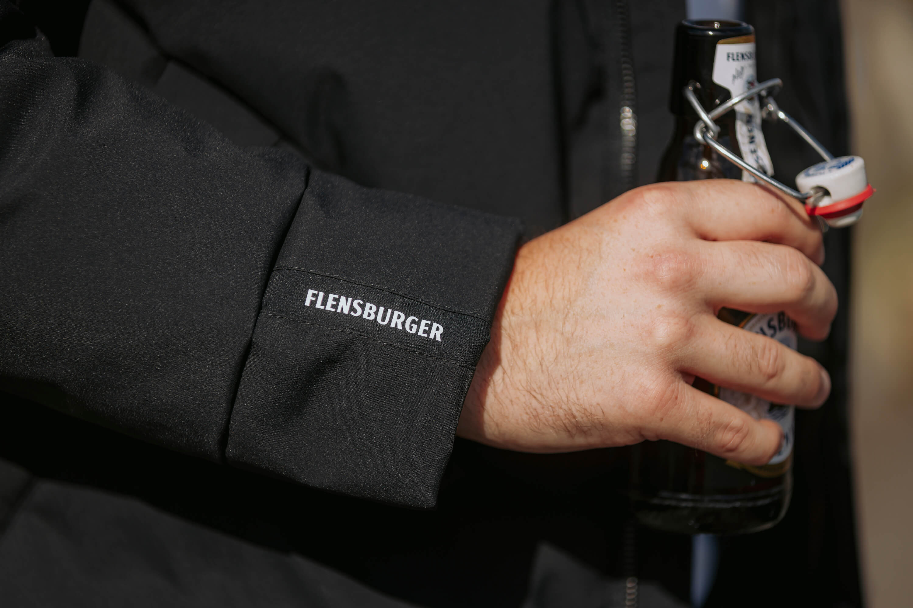 Hand hält eine Flensburger Bierflasche mit Bügelverschluss. Das schwarze Jackenärmel zeigt den weißen Schriftzug „FLENSBURGER“.
