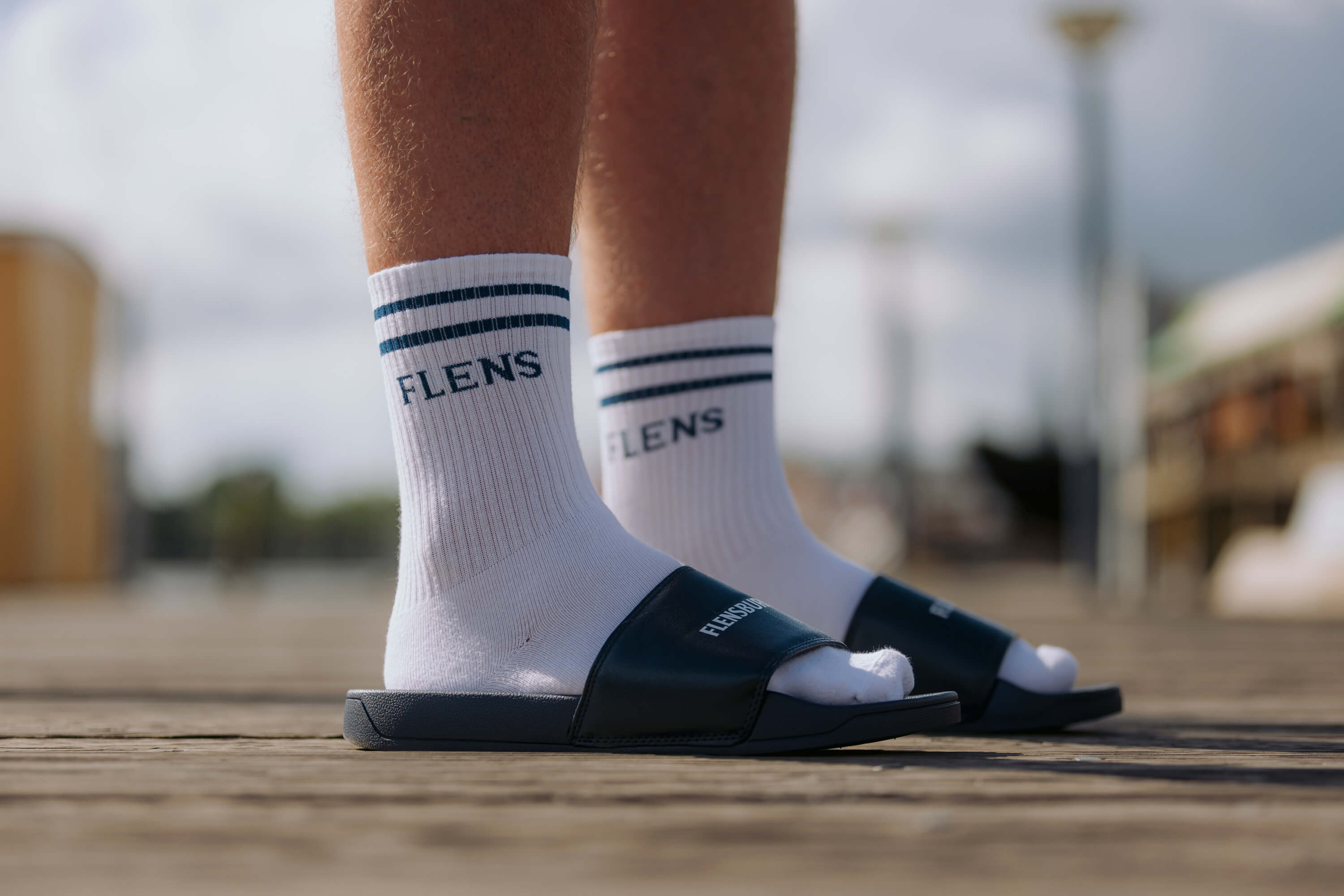 Weiße Sportsocken mit zwei blauen Streifen und dem Schriftzug "FLENS" werden getragen. Die Socken sind mit schwarzen Schlappen mit der Aufschrift "FLENSBURG" kombiniert.