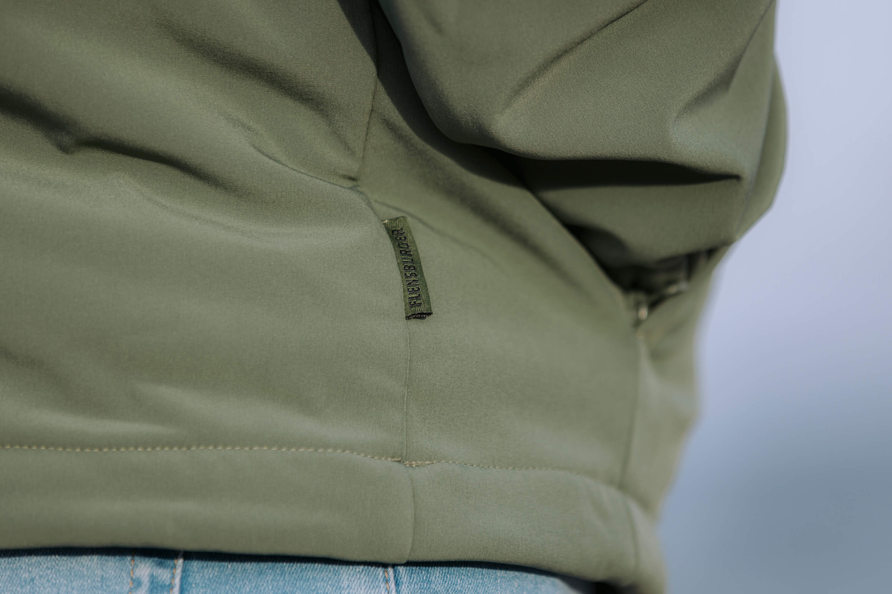 Grüner, wetterfester Windbreaker mit Nahtdetail und schwarzem Textlabel "WINDSBREAKER" am Bündchen. Die Jacke endet auf blauen Jeansstoff.