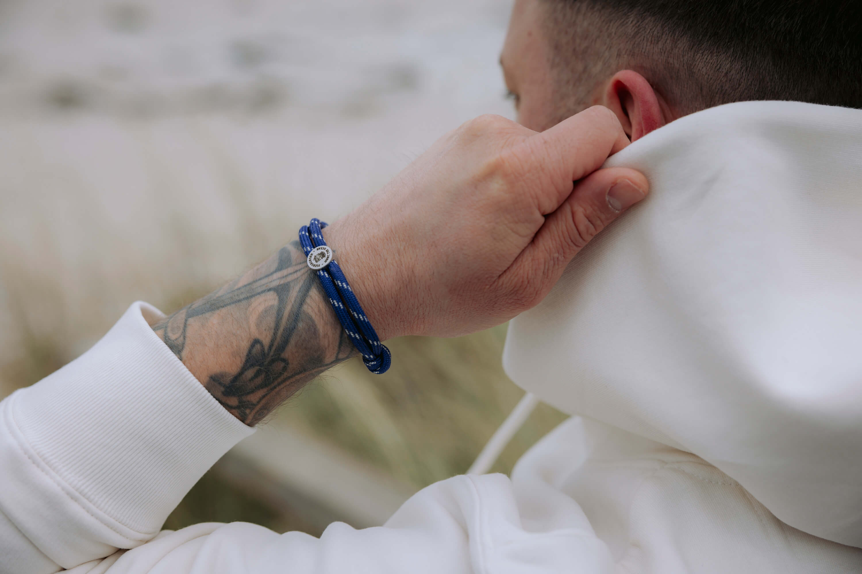 Ein Arm mit einem blauen Armband aus Garn aus 100% rPET in einer weiße Kapuzenjacke. Das Armband hat eine kleine silberne Plakette.