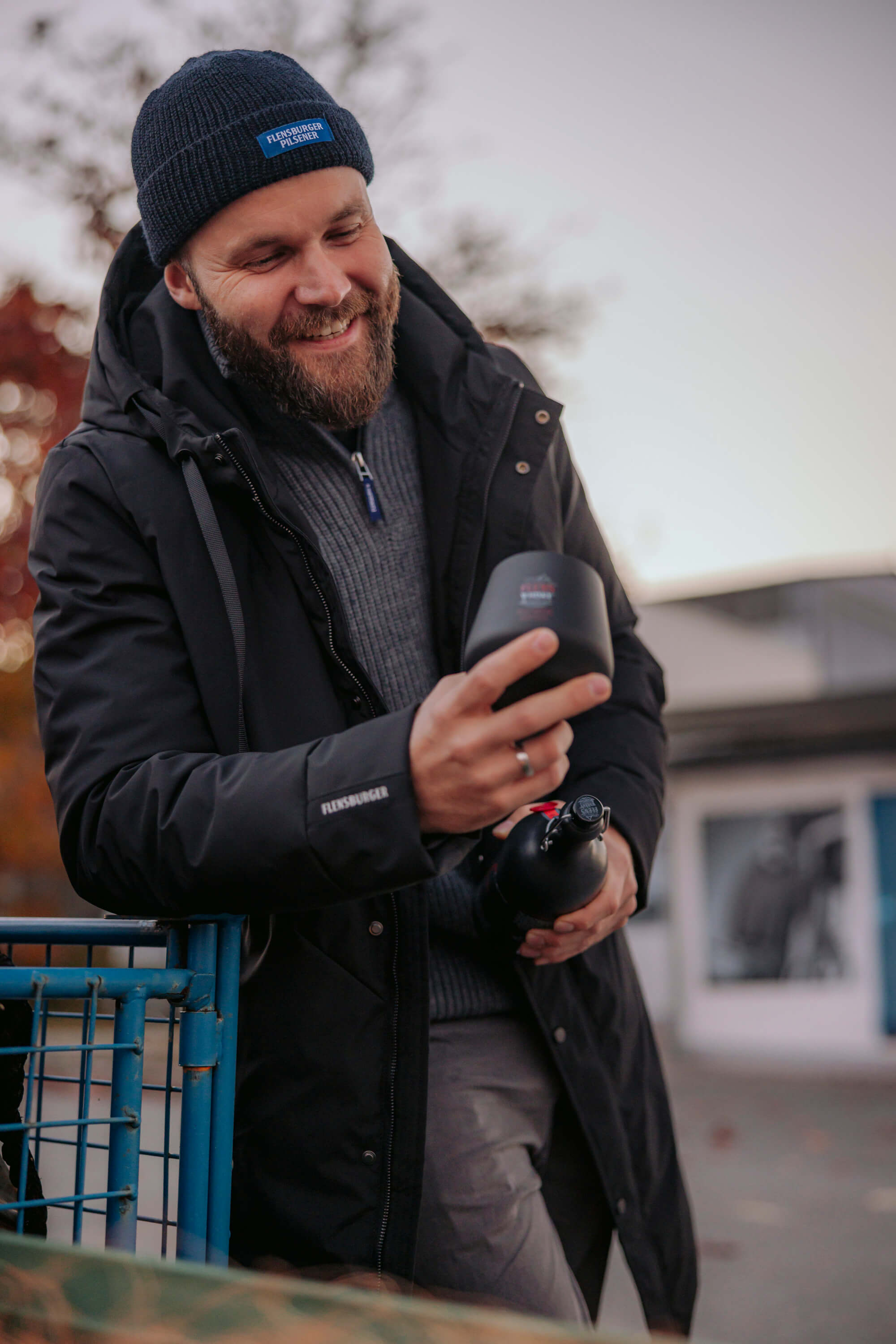 Mann trägt eine schwarze Winterjacke mit dem Schriftzug "Flensburger" auf dem Arm und eine blaue Mütze mit dem Schriftzug "FLENSBURGER PILSENER". Er hält eine schwarze Flasche und einen schwarzen Becher, lächelt dabei.