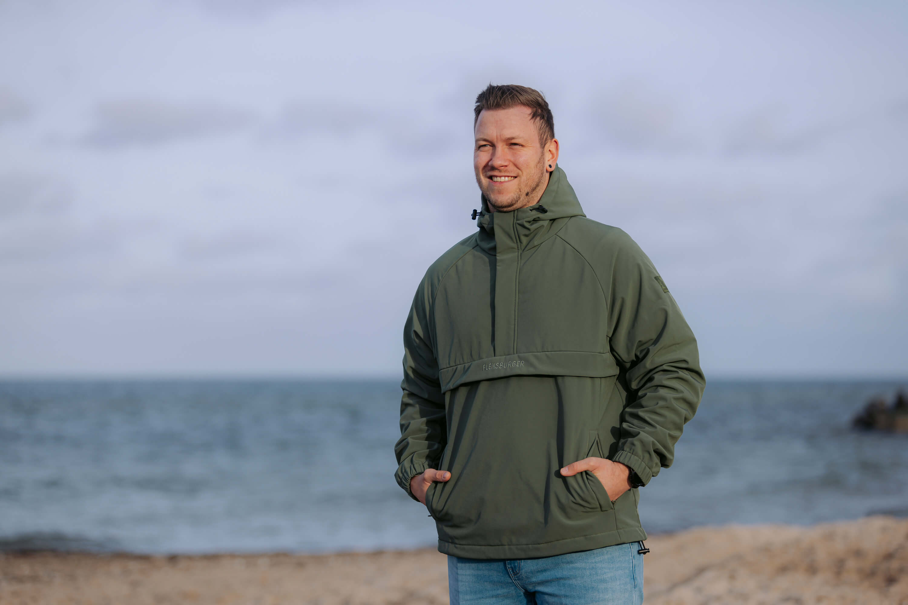 Ein Mann trägt den wetterfesten Windbreaker "Skagerrak". Er steht mit den Händen in den Taschen am Strand vor dem Meer.