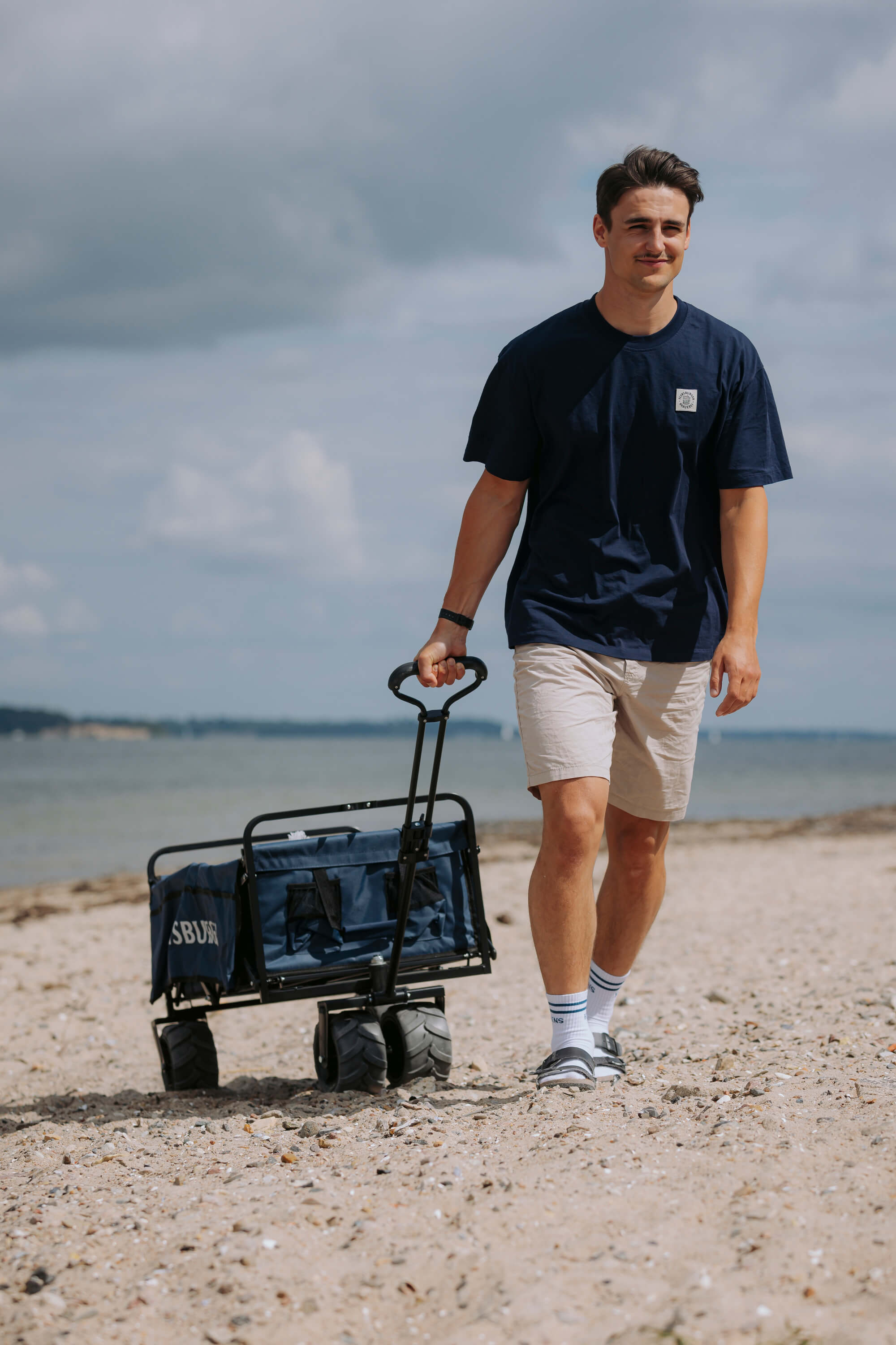 Ein junger Mann zieht den faltbaren Bollerwagen "Rasmus" mit großen Reifen am Strand entlang. Er trägt ein dunkelblaues T-Shirt, helle Shorts sowie weiße Socken mit Sandalen.