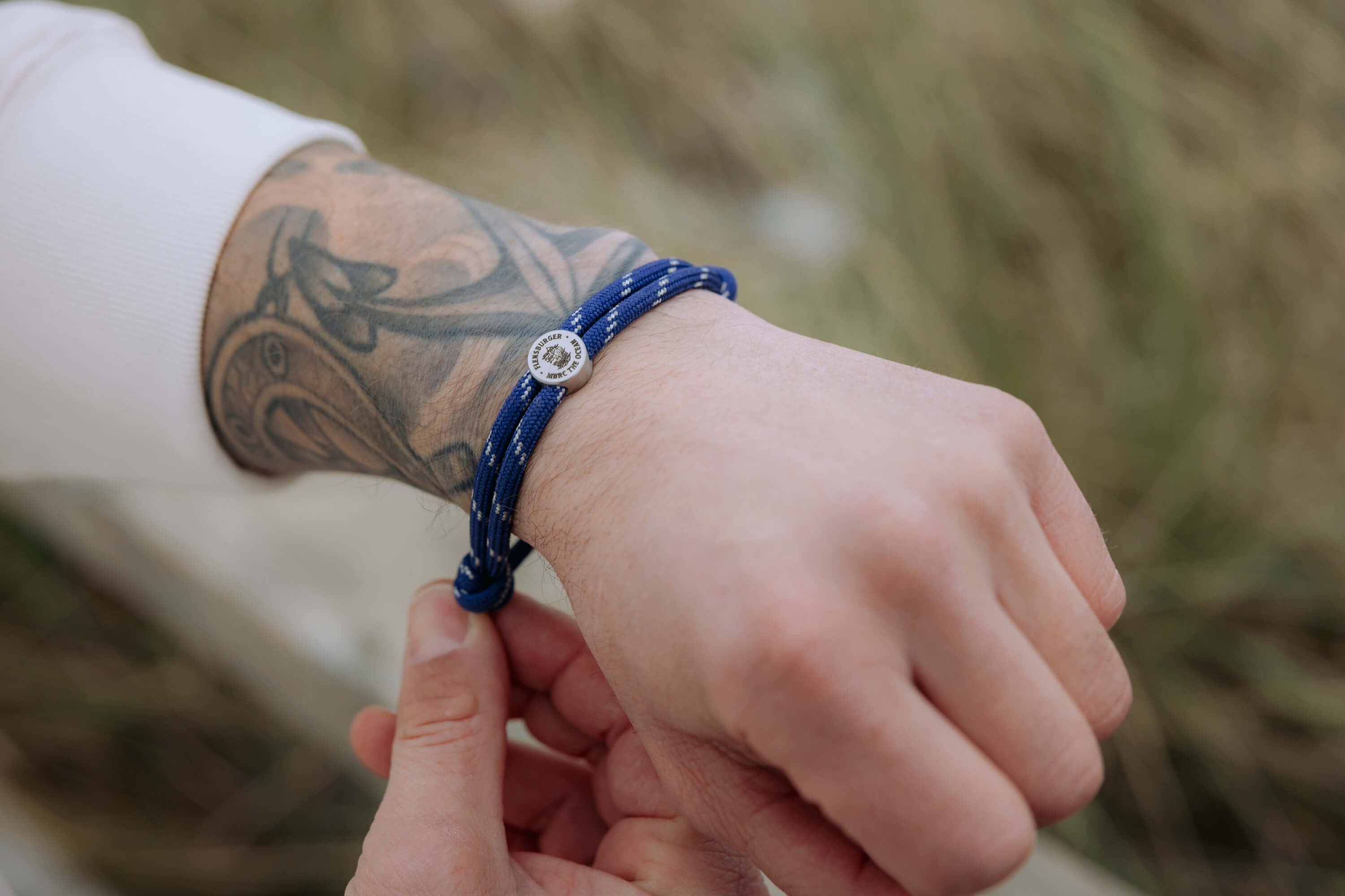 Ein tätowierter Arm trägt ein blaues Armband aus recyceltem Material mit runden Metallplakette und dem Schriftzug „FLENSBURGER MBRC TM 2022“. Eine Hand hält das Armband.