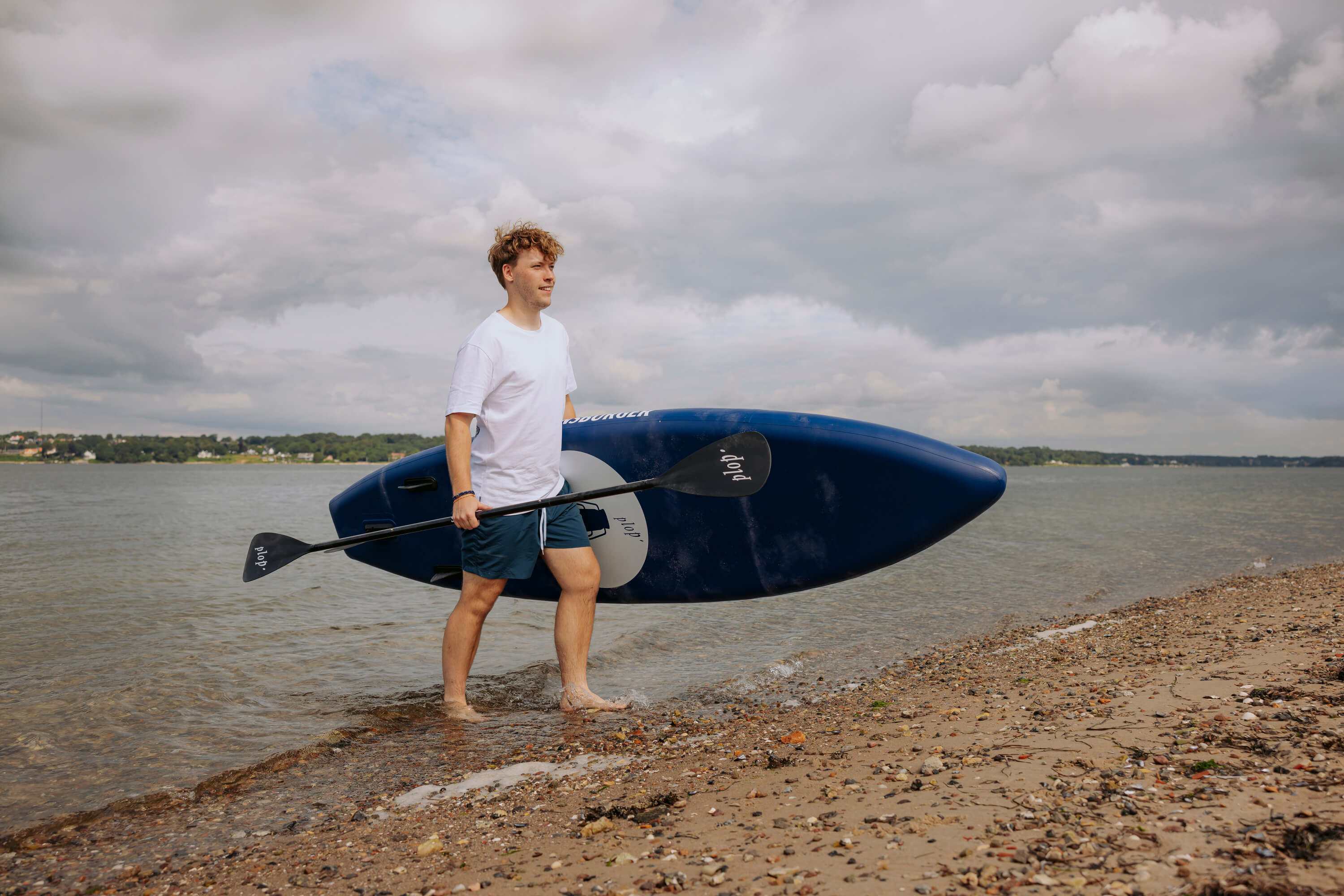 Mann trägt ein blaues SUP Board und ein Paddel am Strand, steht barfuß am Wasser, trägt weiße T-Shirt und blaue Shorts, bewölkter Himmel im Hintergrund.