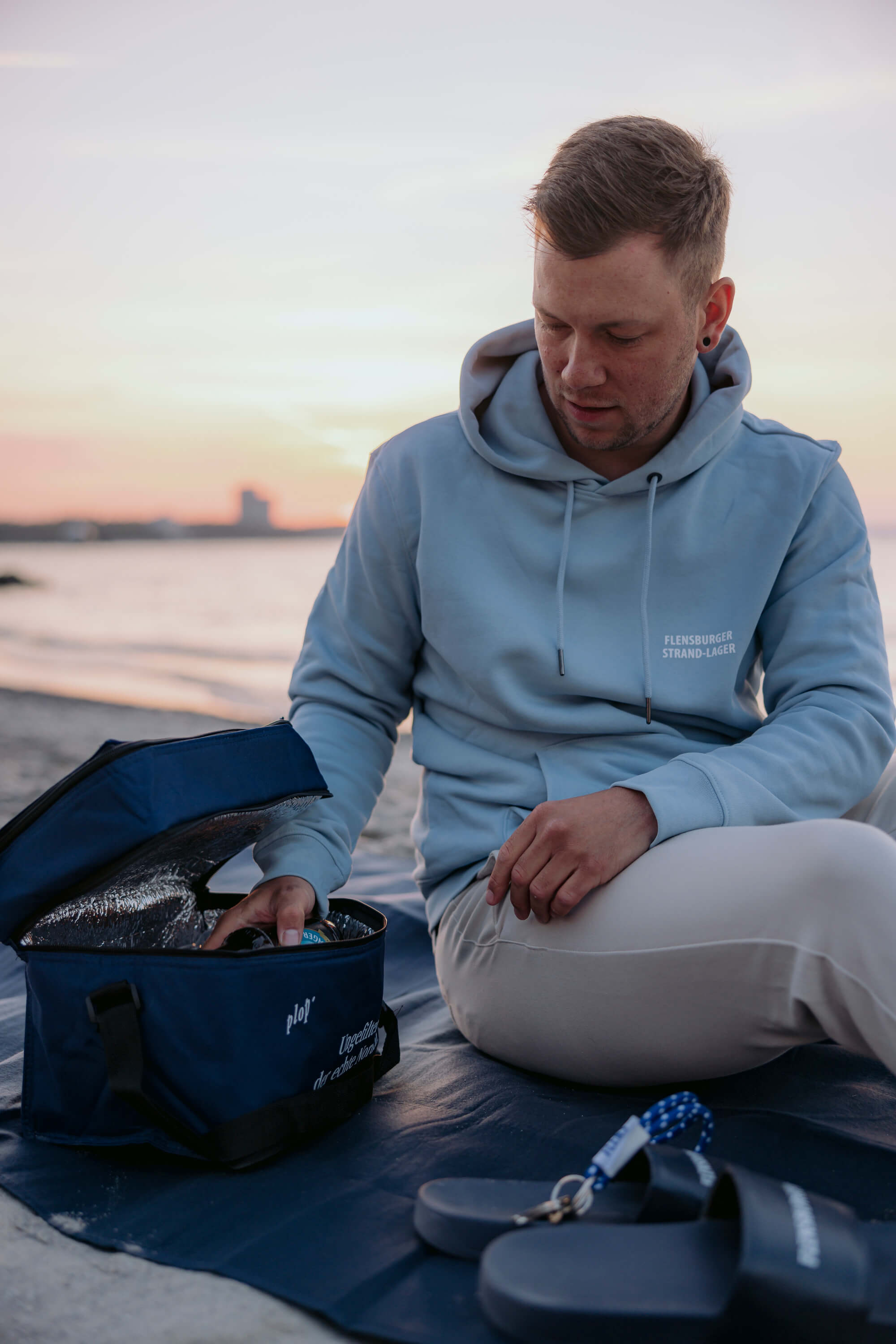 Mann in hellblauem Hoodie sitzt am Strand und öffnet eine blaue Kühltasche. Er prüft den Inhalt der Tasche, während die Sonne untergeht.