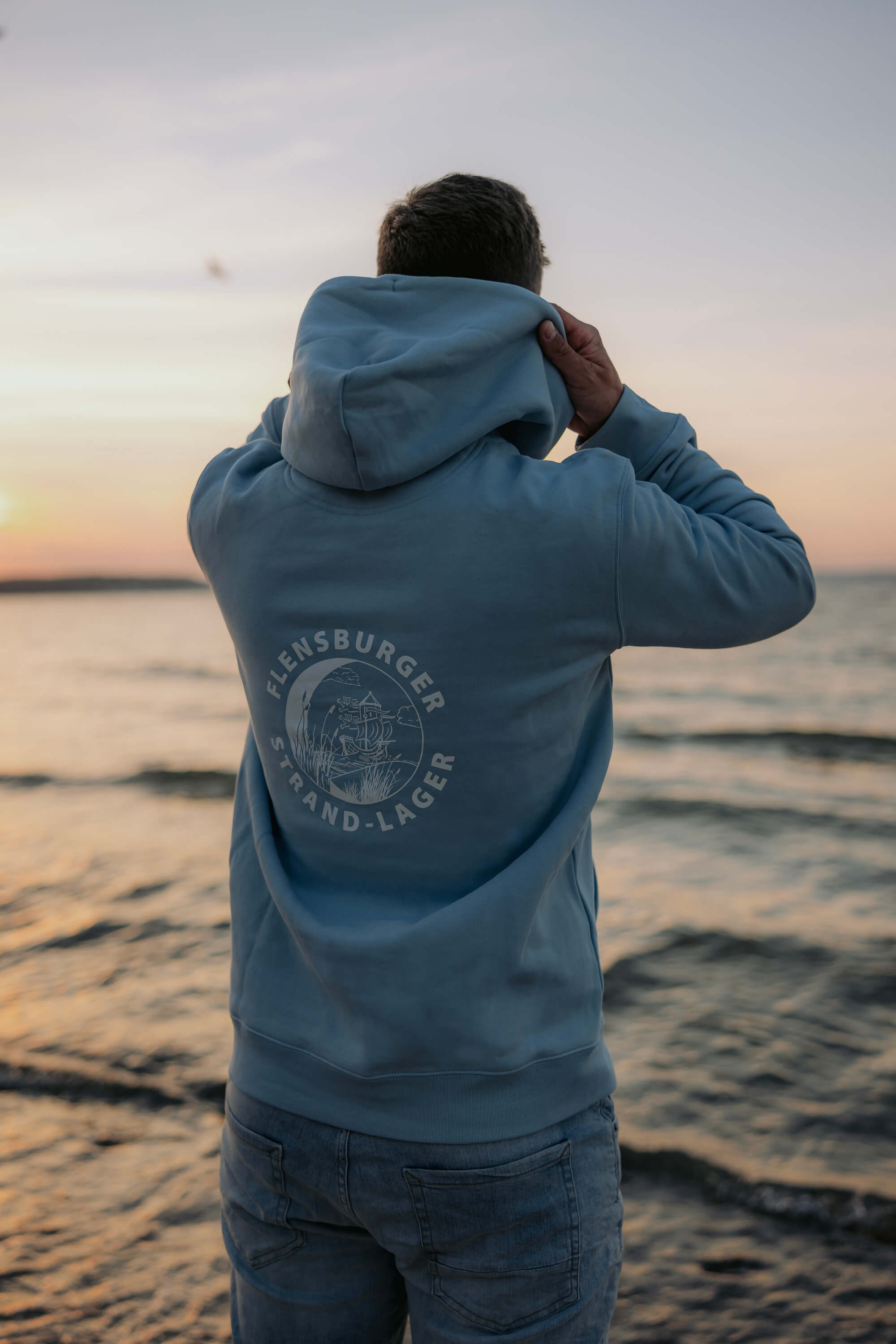Ein Mann trägt einen hellblauen Hoodie mit der Aufschrift „Flensburger Strand-Lager“ und einer Schiffsgrafik auf dem Rücken. Er steht am Ufer und blickt auf das Wasser bei Sonnenuntergang.