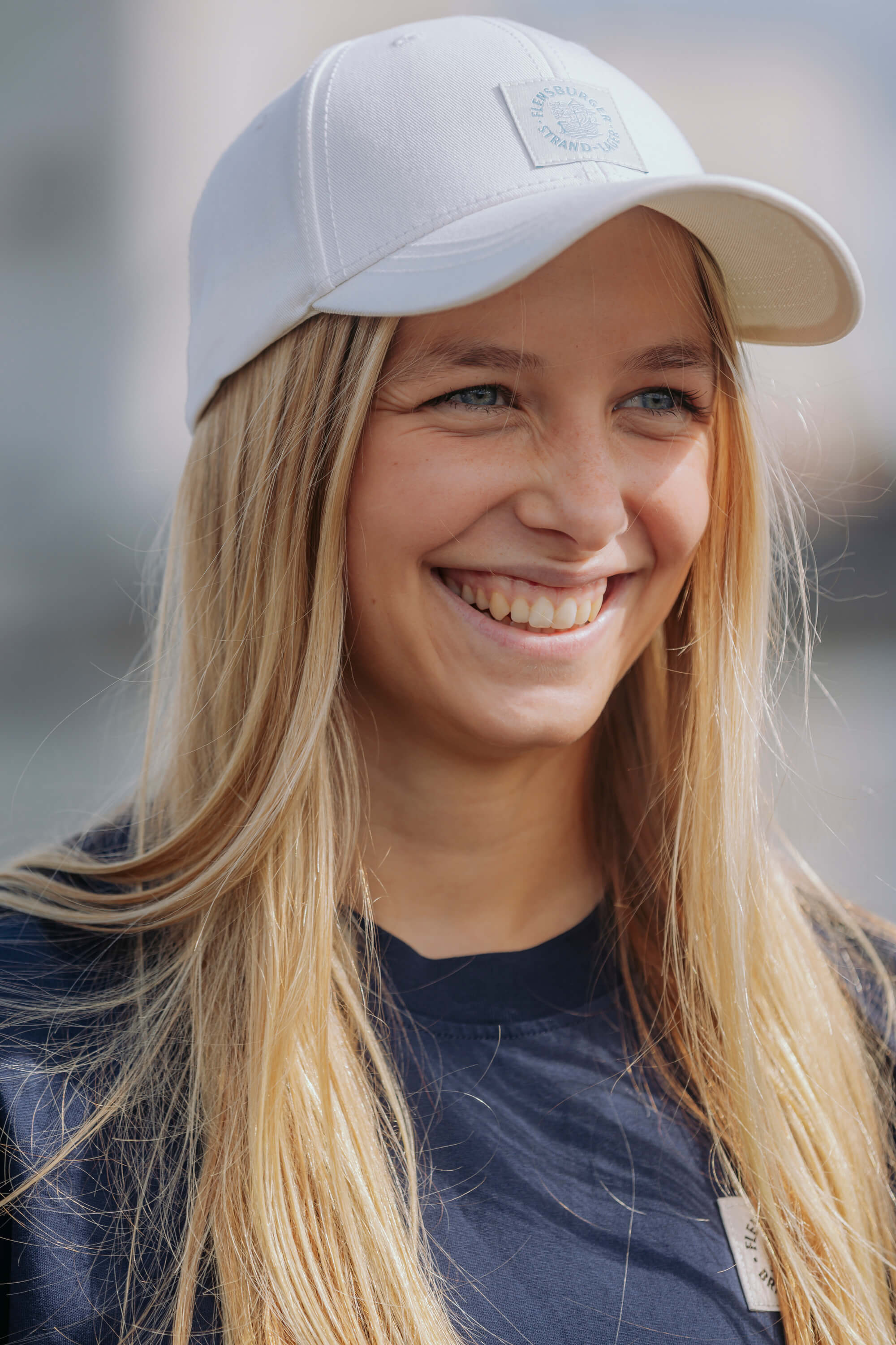 Junge lächelnde Frau trägt die strandbeige Strand-Lager Cap von FLENS mit sichtbarem Weblabel. Sie hat lange blonde Haare und ein dunkelblaues Shirt.