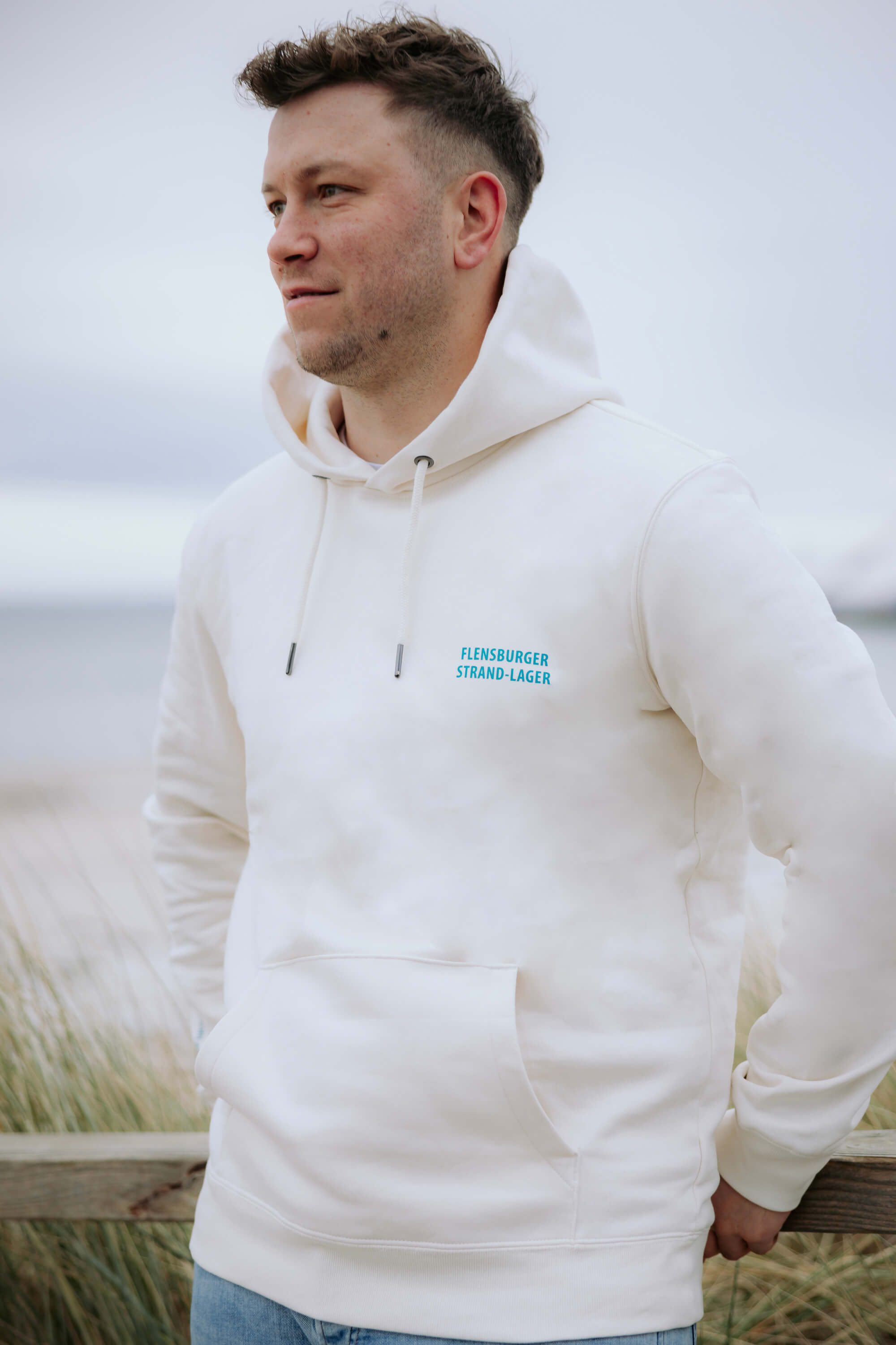 Ein Mann trägt einen beigen Hoodie mit der blauen Aufschrift „FLENSBURGER STRAND-LAGER“. Er lehnt entspannt an einem Holzgeländer am Strand.
