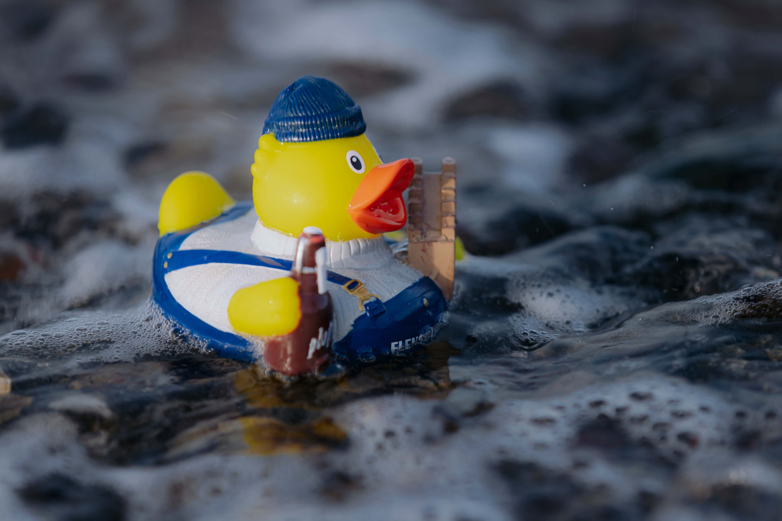 Die gelbe Gummiente trägt eine blaue Mütze und Latzhose. Sie hält eine braune Flasche und ein braunes Objekt. Die Ente schwimmt im Wasser mit Bläschen.