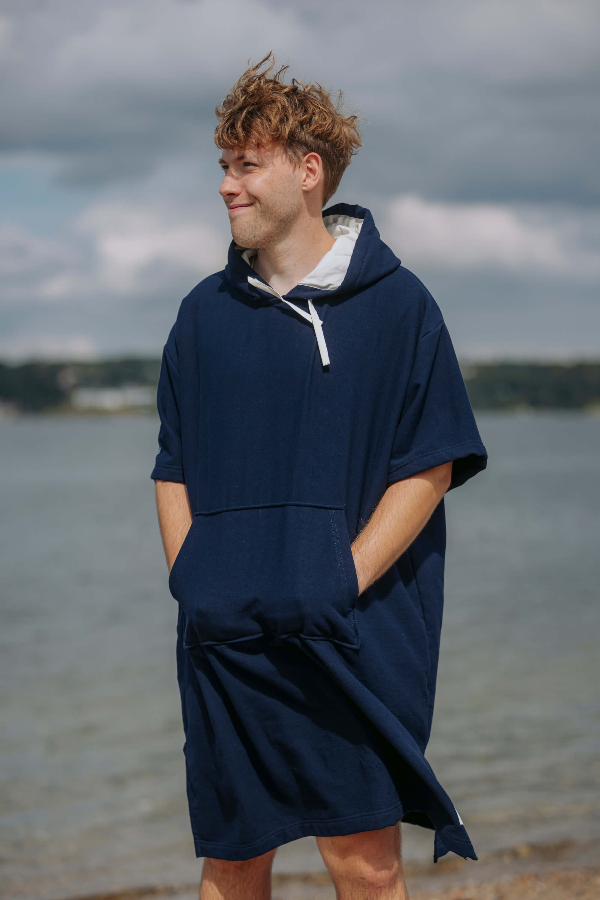 Junger Mann trägt den blauen Strand-Poncho mit weißen Akzenten. Er hat kurze Ärmel und große Taschen vorne. Im Hintergrund ist ein See mit bewölktem Himmel zu sehen.