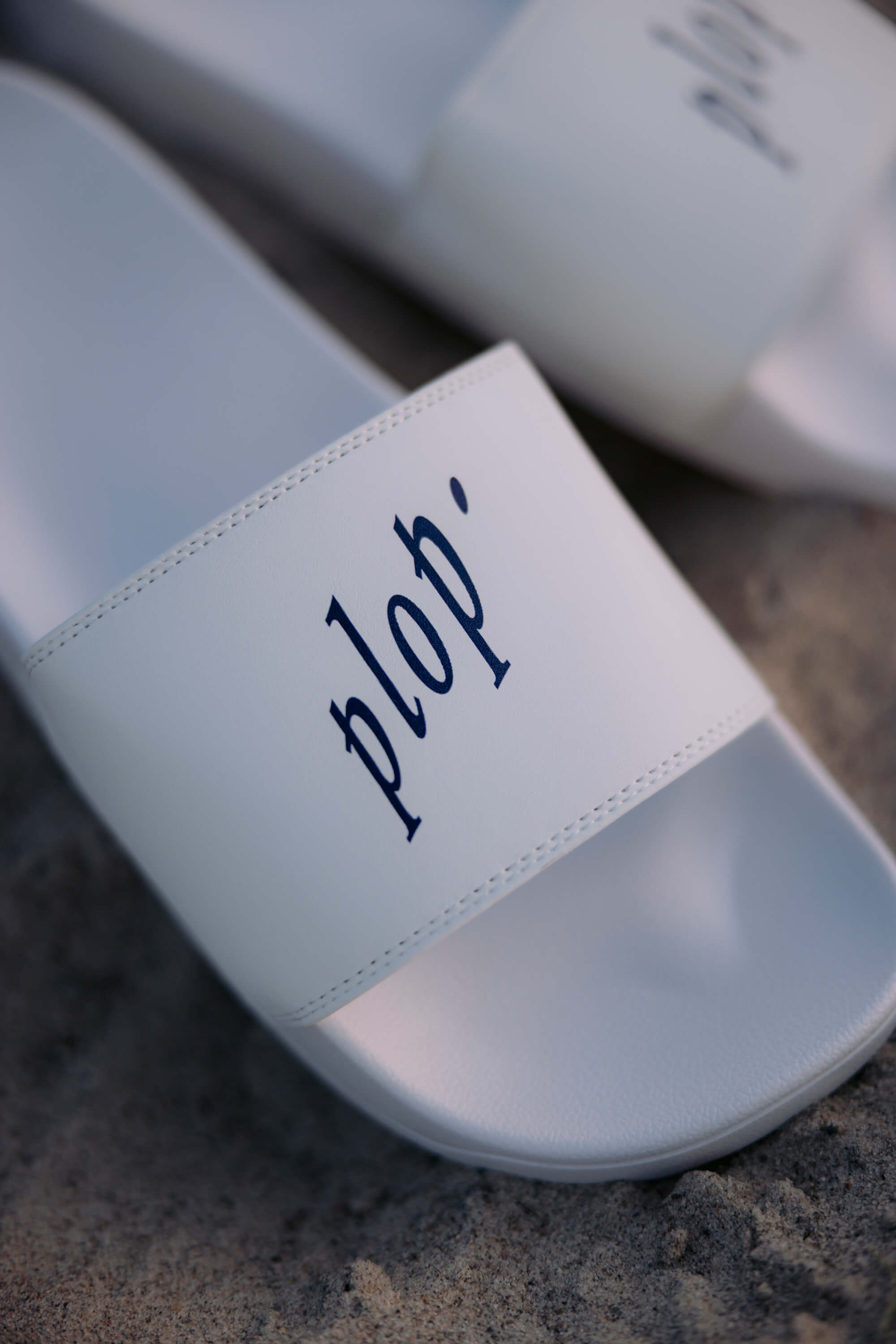 Weiße Fördeslipper liegen auf Sand. Auf dem Riemen steht in blauer Schrift „plop'“.