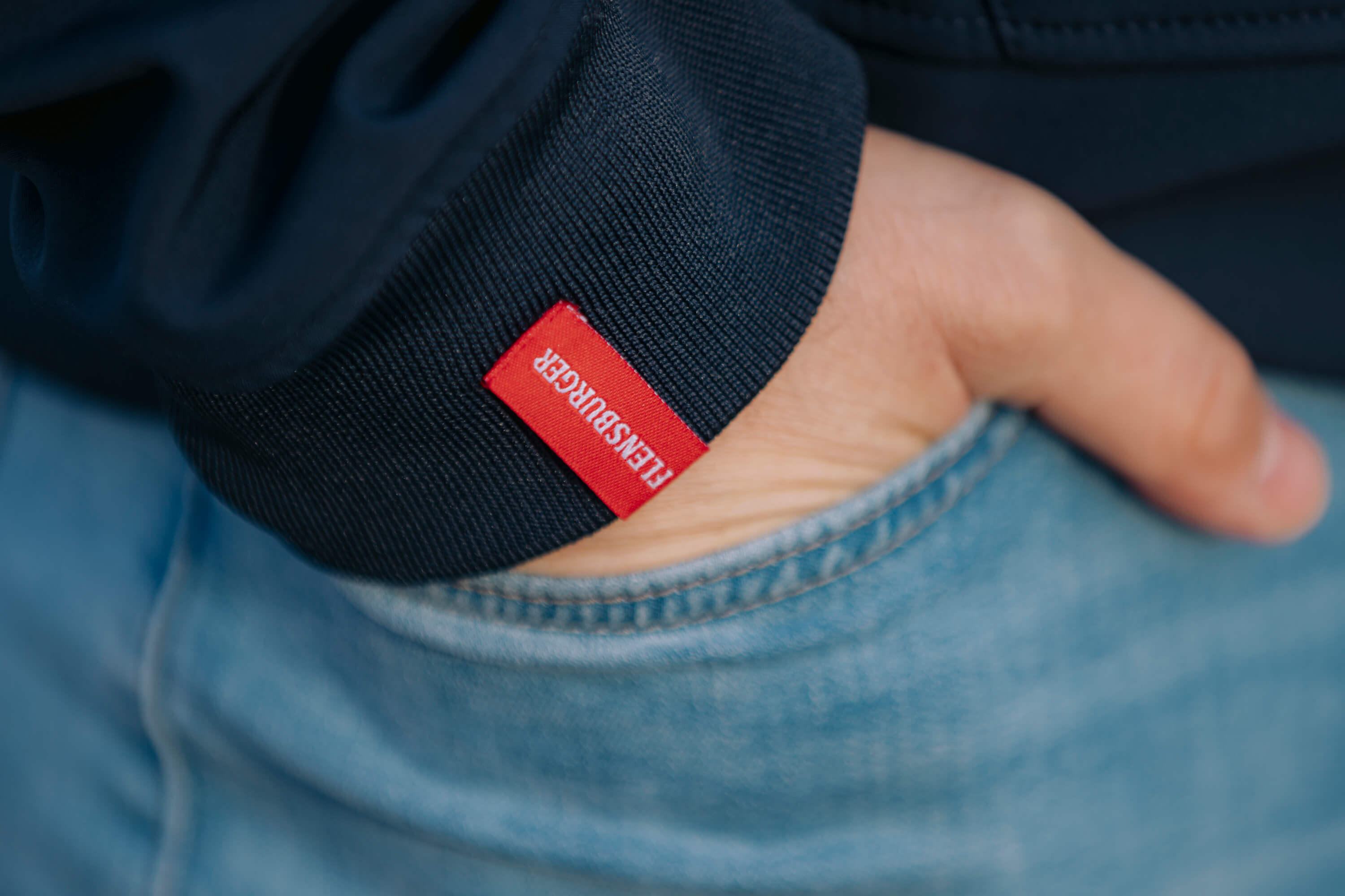 Eine Hand steckt in der Hosentasche einer blauen Jeans. Der Ärmel einer dunkelblauen Softshelljacke mit rotem Etikett "FLENSBURGER" ist sichtbar.