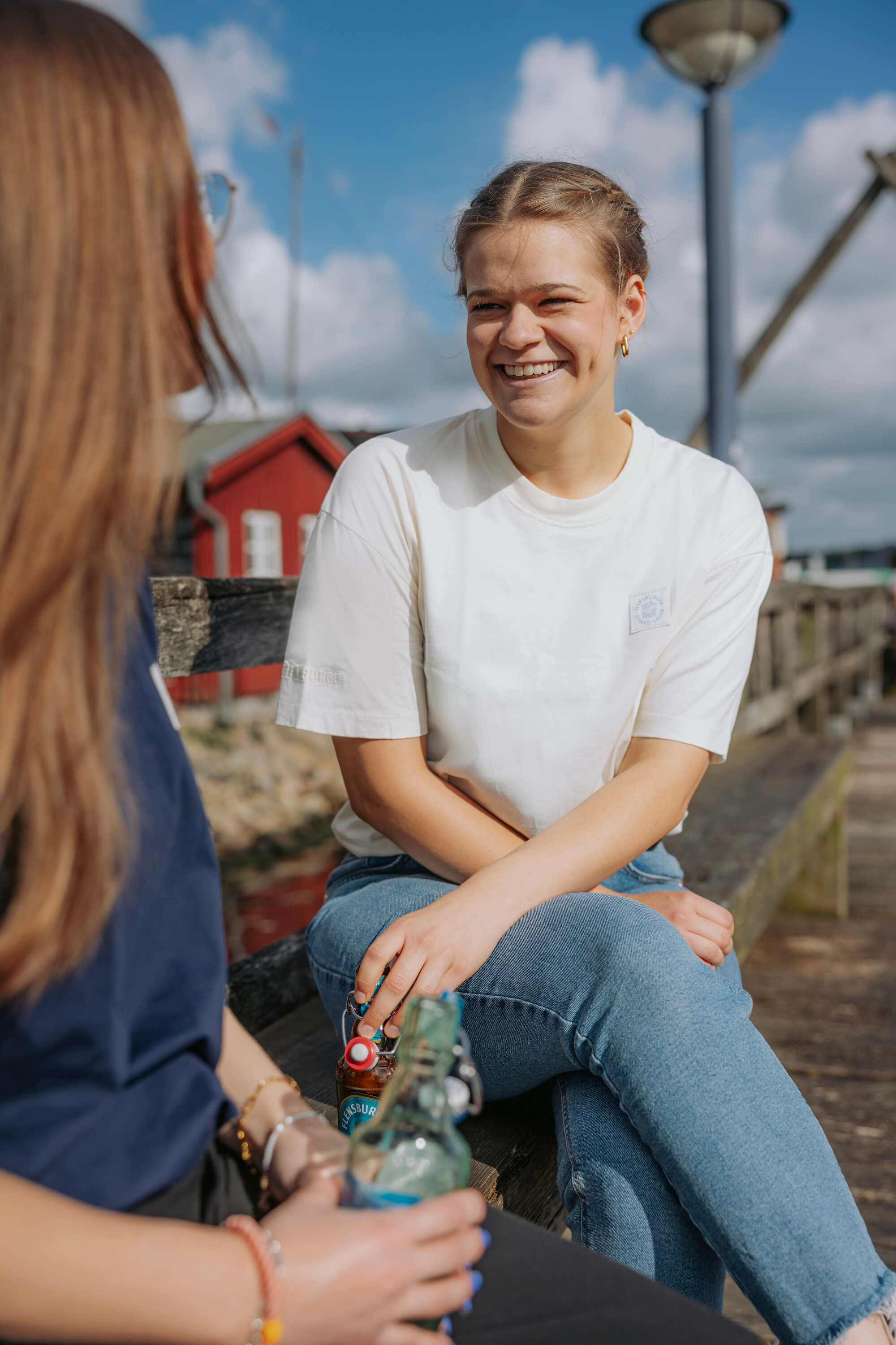 Zwei Frauen sitzen auf einer Holzbank im Freien und unterhalten sich. Die Frau rechts trägt ein beige Kurzarmshirt und blaue Jeans, die andere hält eine Flasche.