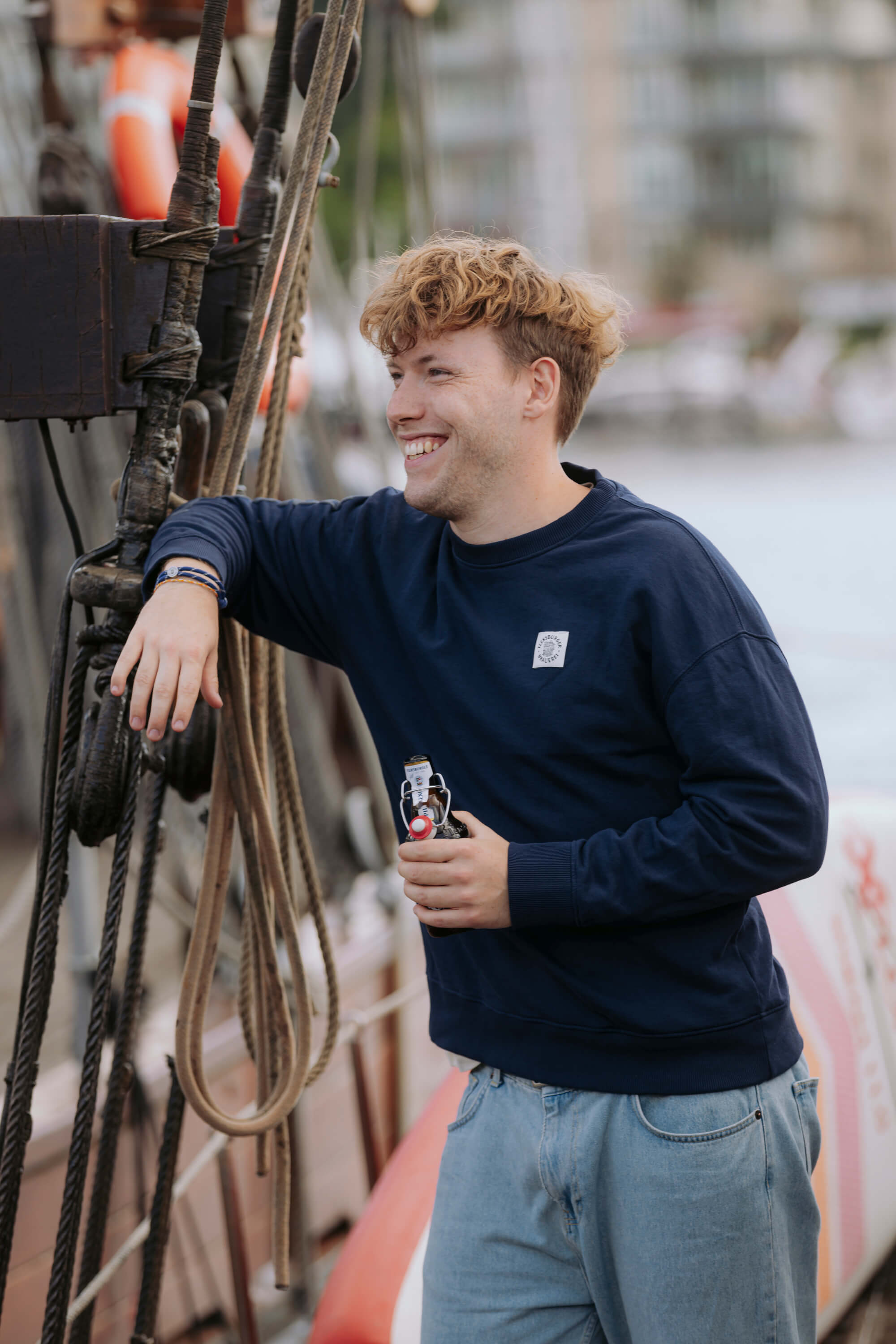 Junger Mann lehnt lächelnd an einem Schiffsmast und hält eine Flasche in der Hand. Er trägt einen dunkelblauen Basic Sweater mit einem kleinen Weblabel auf Herzhöhe.