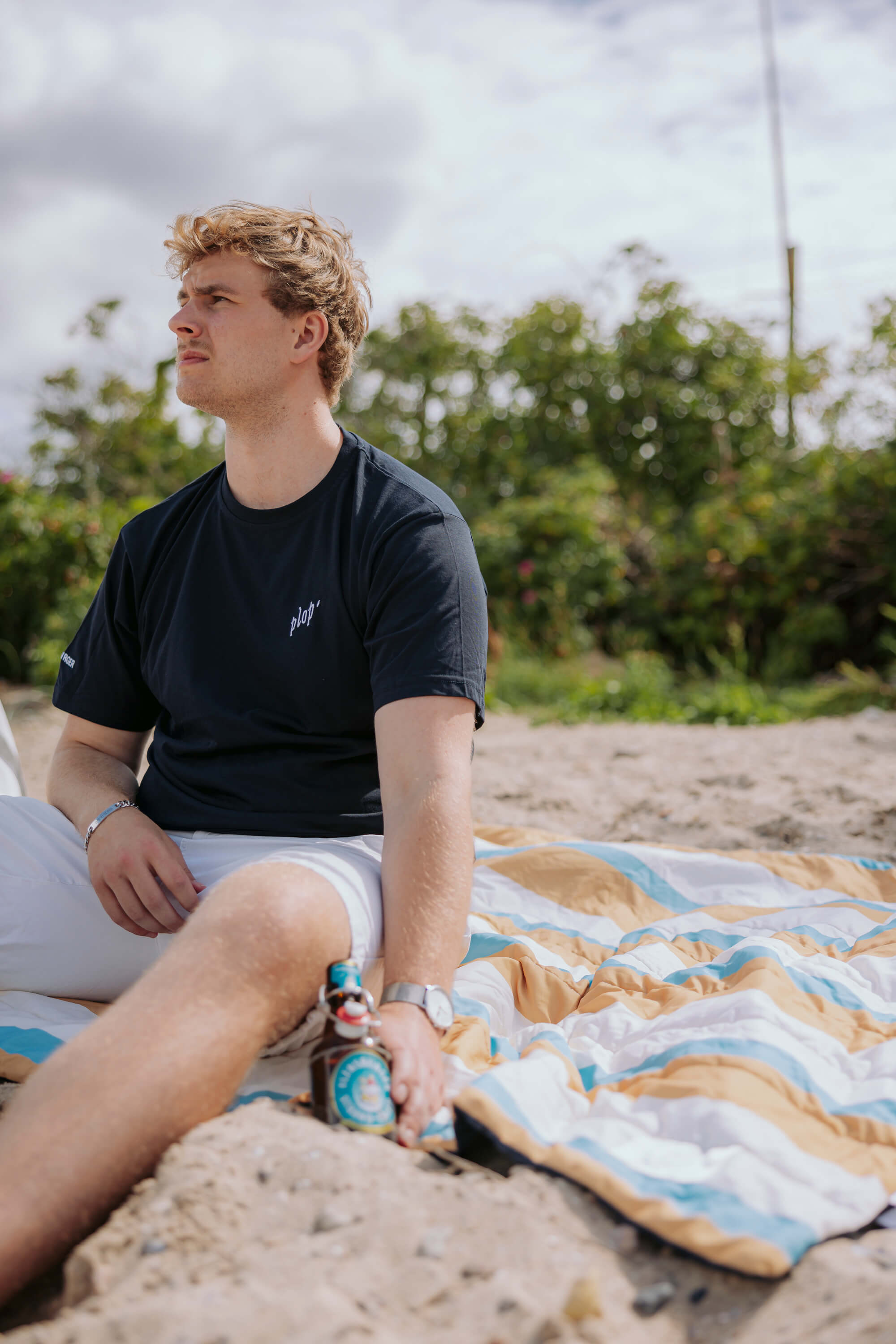 Junger Mann sitzt auf einer 3in1 Outdoordecke mit Petrol-Blau-Beige-Braunen Streifen. Er trägt ein schwarzes T-Shirt mit weißem Schriftzug "plop" und weiße Shorts.
