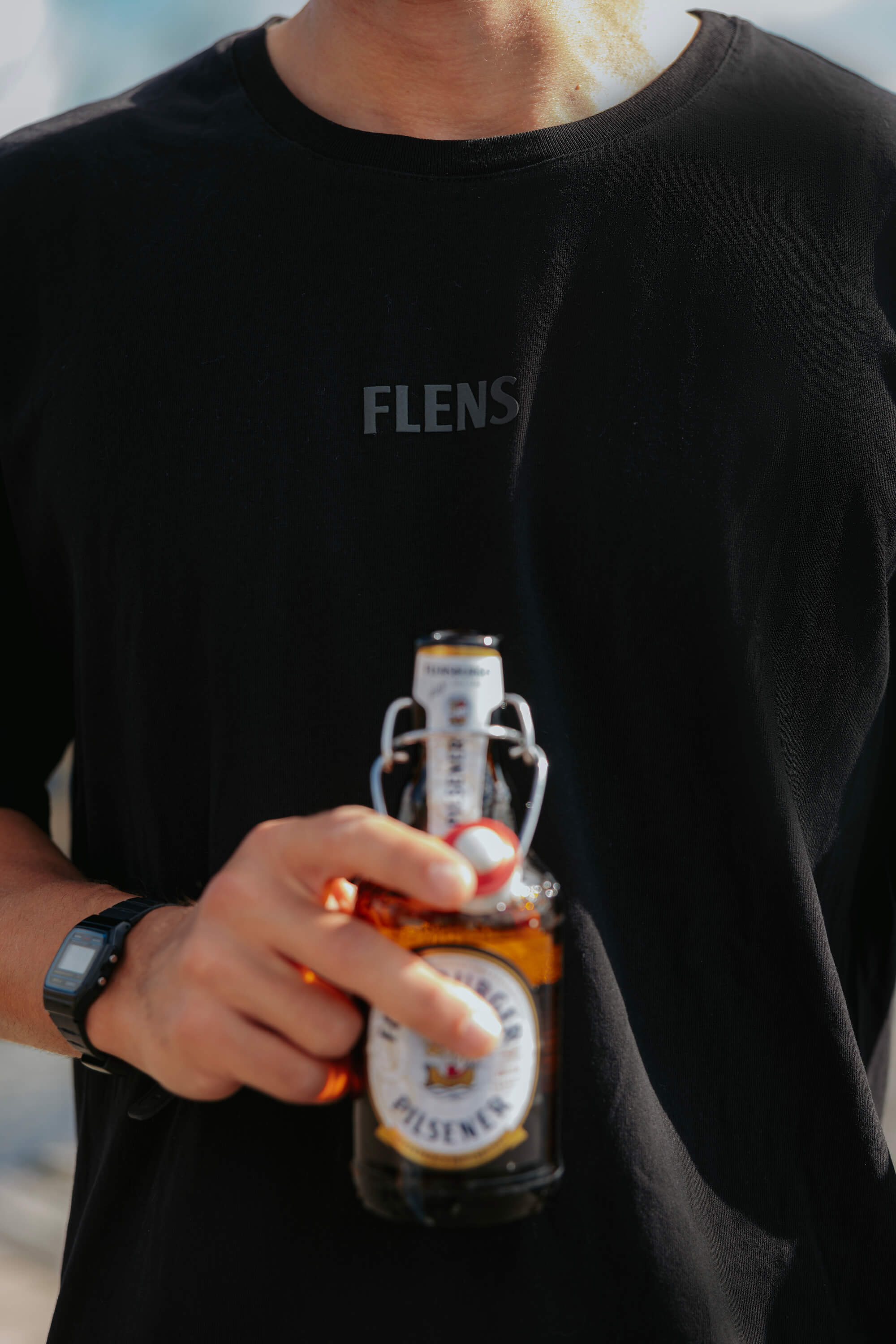 Person trägt ein anthrazit-schwarzes Shirt mit der Aufschrift "FLENS" auf der Brust. Die Person hält eine Flasche Flensburger Pilsener Bier mit Bügelverschluss in der Hand.