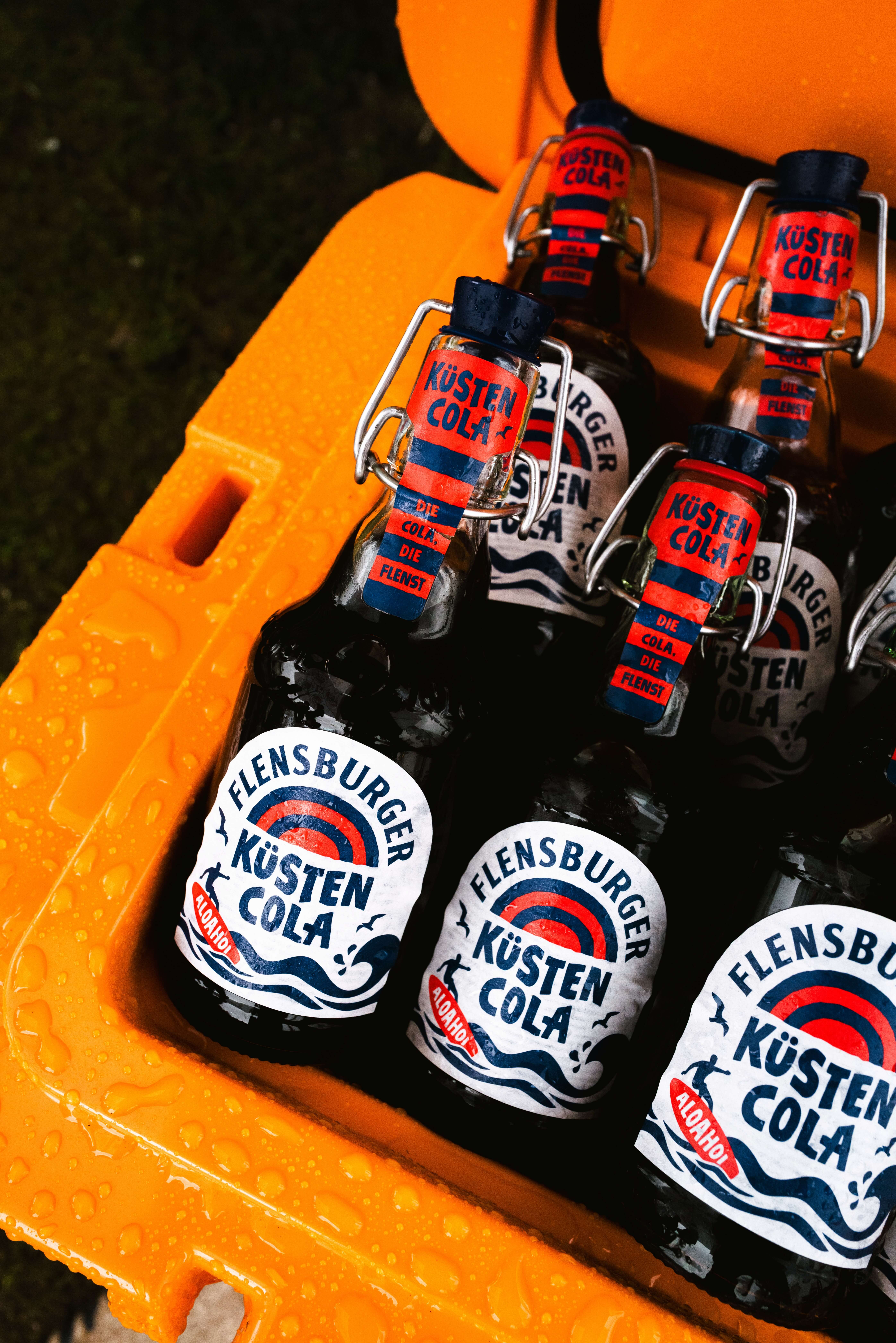 Sechs Flensburger Küstencola-Flaschen in einer orangefarbenen Kiste. Die Flaschen besitzen einen Bügelverschluss und sind mit einem Etikett versehen, das den Namen und die Beschreibung der Cola anzeigt.