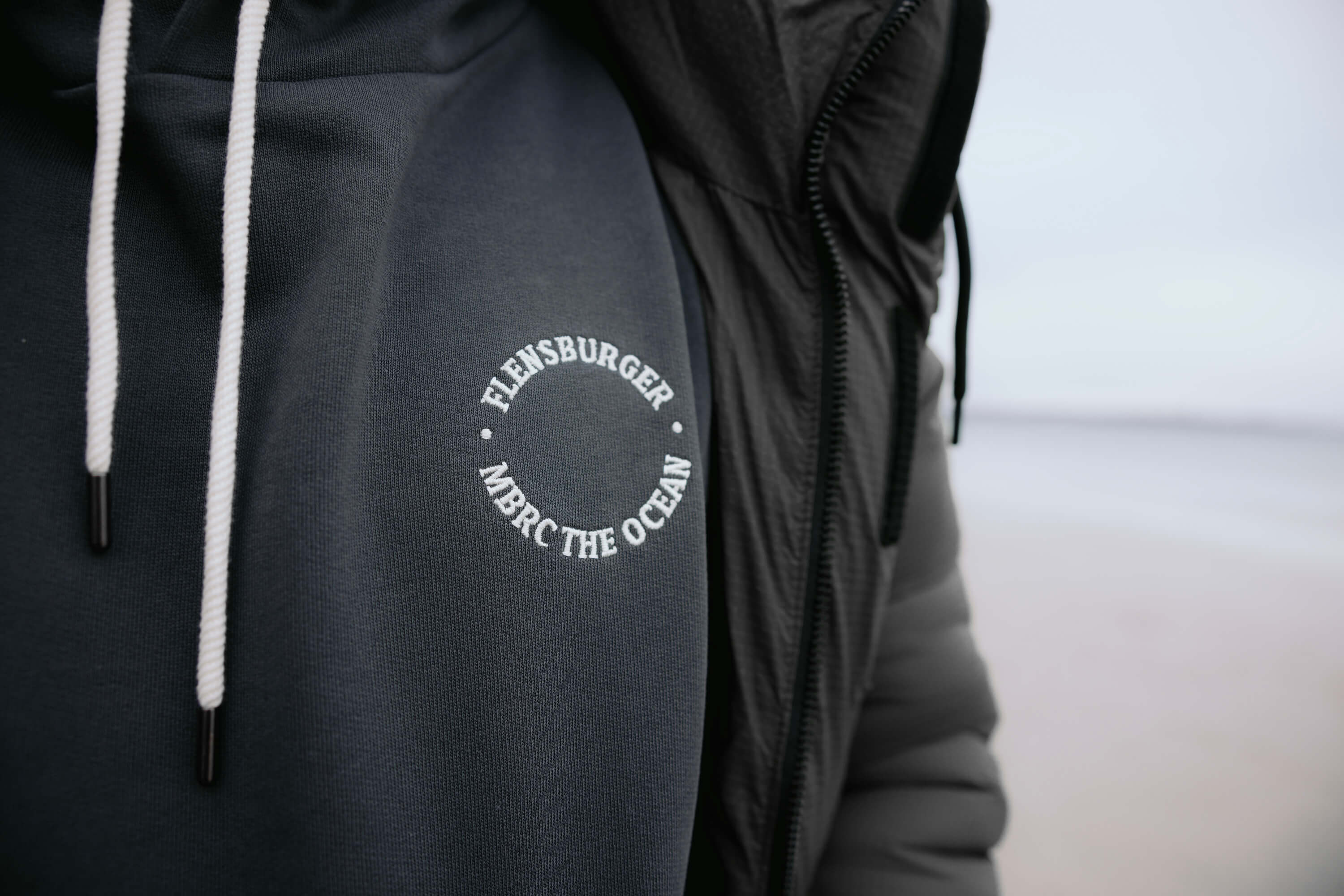 Hoodie in Anthrazit mit weißem Schriftzug "FLENSBURGER MBRC THE OCEAN" in kreisförmiger Anordnung auf der linken Brust. Der Hoodie hat weiße Kordeln am Kragen.