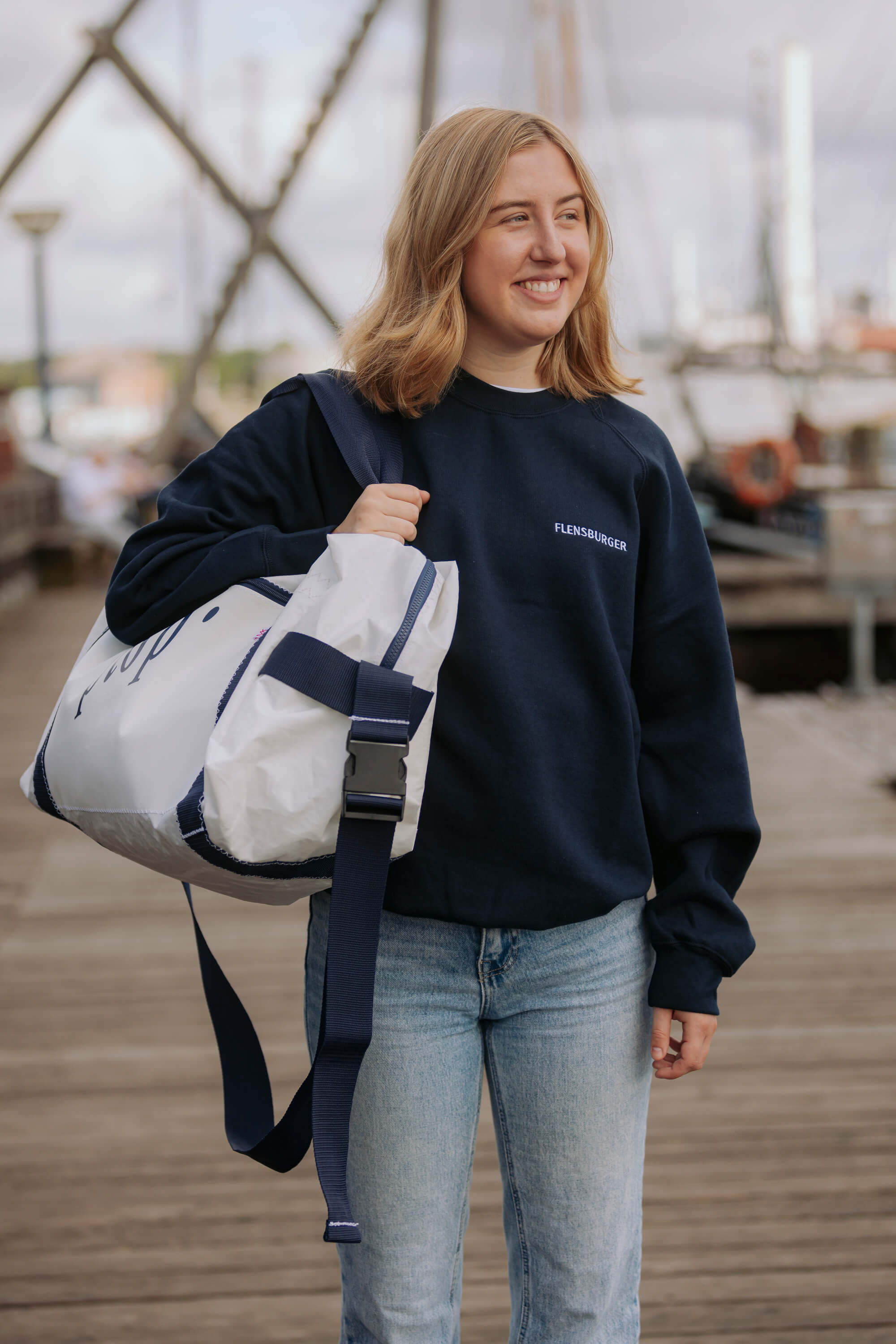 Eine junge Frau trägt den dunkelblauen Pullover mit weißem Aufdruck "FLENSBURGER" auf der Vorderseite. Sie hält eine weiße Tasche mit dunkelblauem Gurt über der Schulter.