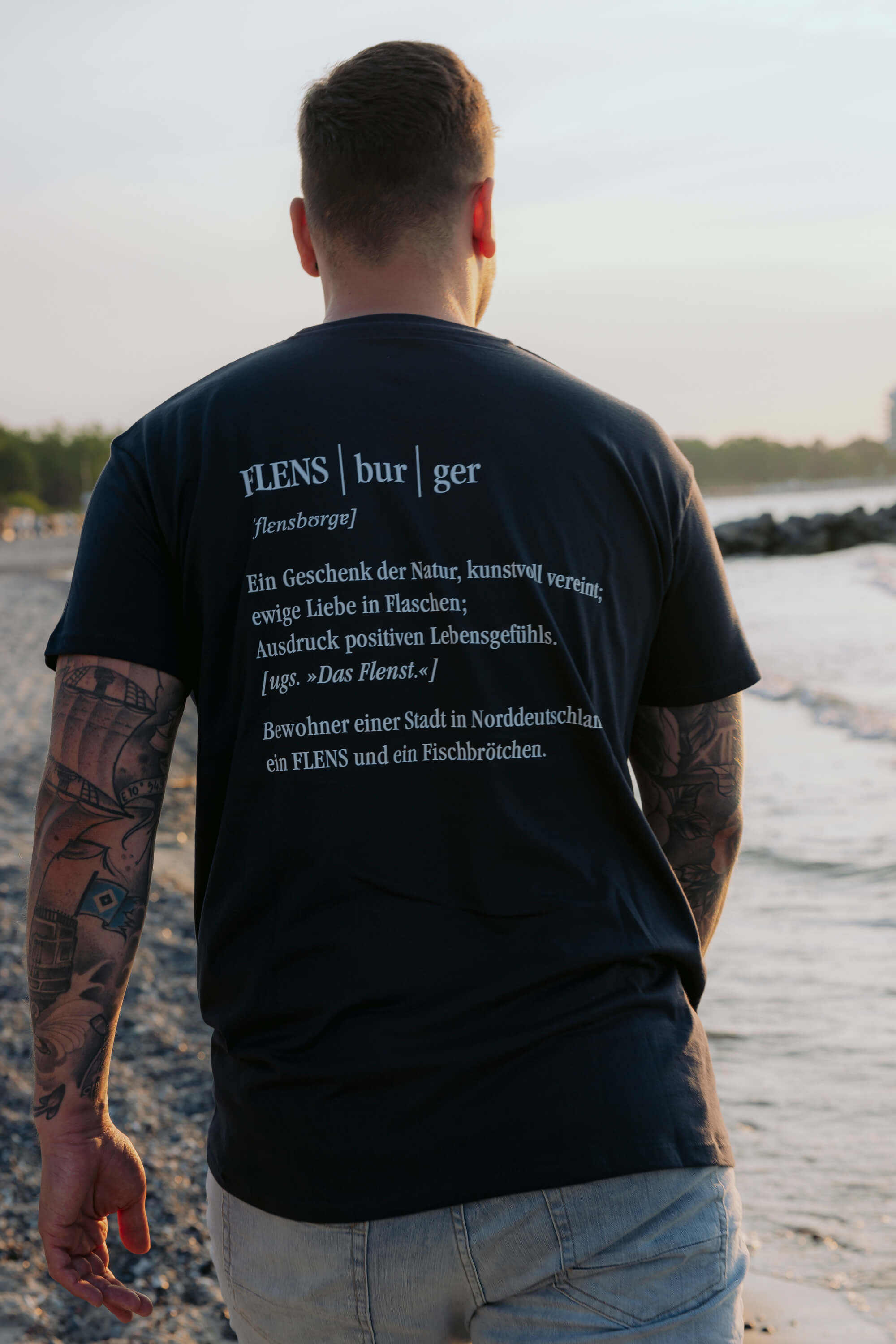 Ein Mann steht am Strand und trägt ein navyblaues T-Shirt mit dem Text „FLENS|bur|ger“ und einer poetischen Beschreibung über Flensburg auf dem Rücken.