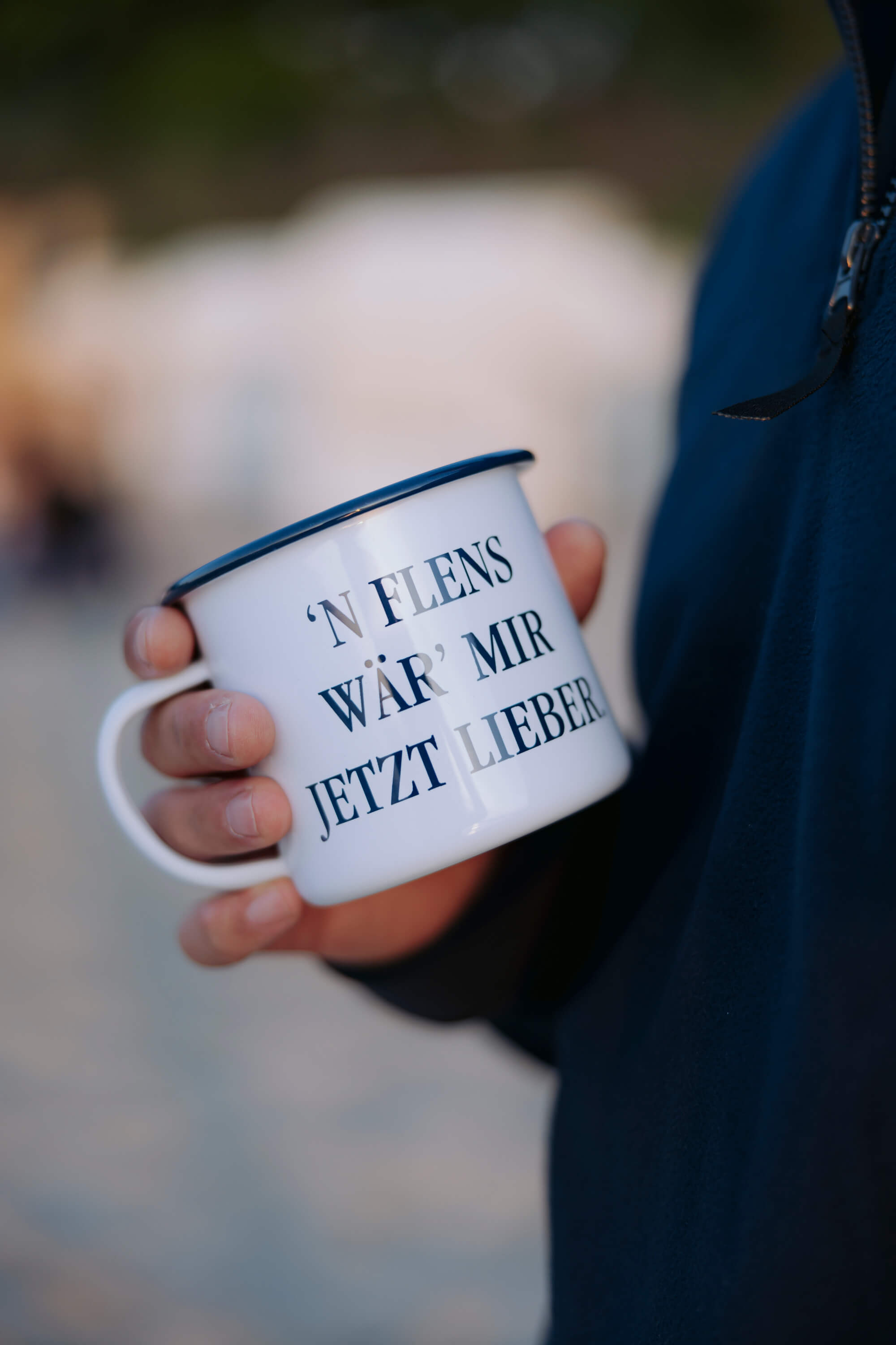 Weiße Emaille-Tasse mit blauem Rand wird von einer Hand gehalten. Auf der Tasse steht in schwarzer Schrift: "'N FLENS WÄR' MIR JETZT LIEBER".