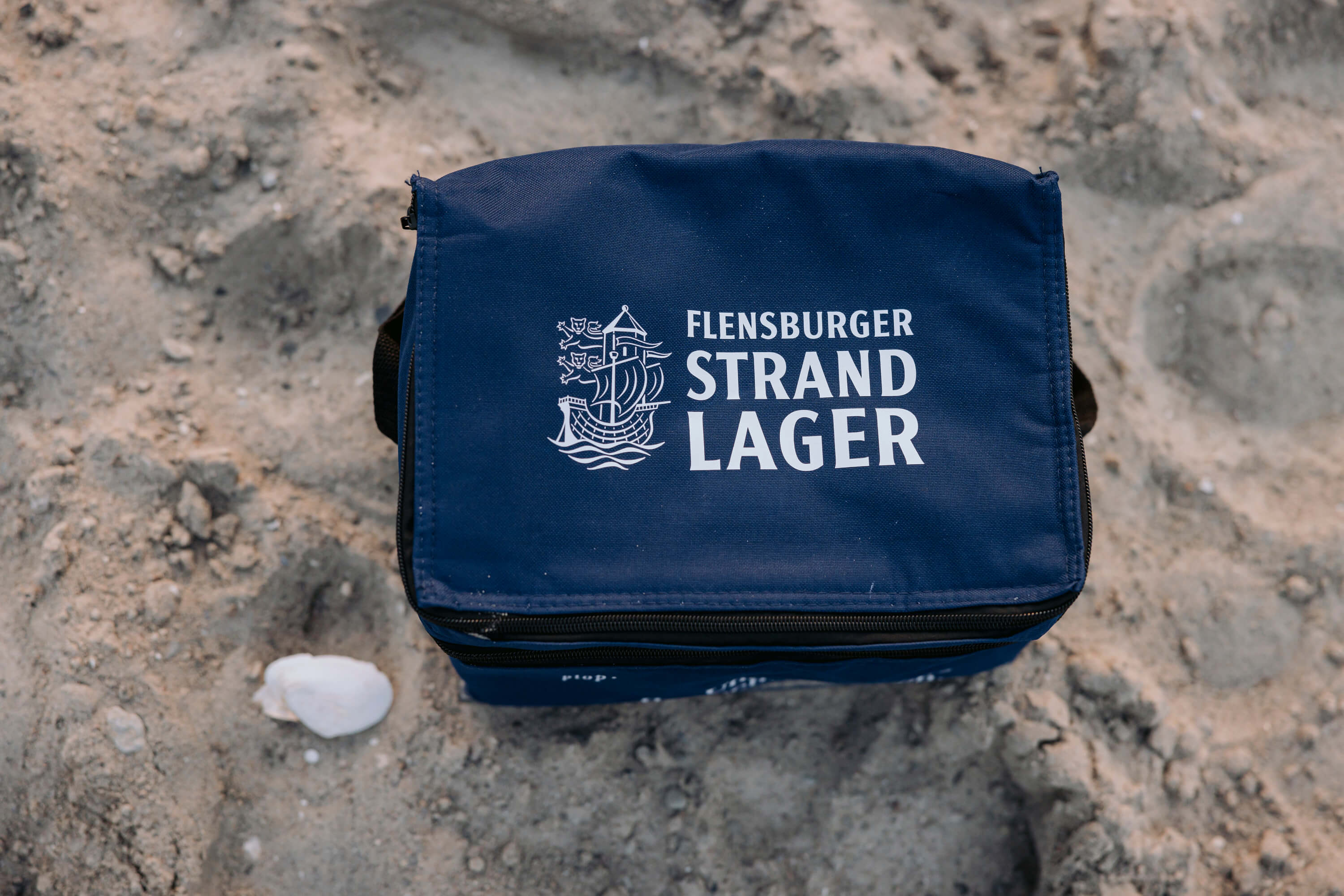Kühlbox in navy blauer Farbe mit dem Aufdruck "FLENSBURGER STRAND LAGER". Die Kühltasche liegt auf sandiger Oberfläche, daneben liegt eine kleine Muschel.
