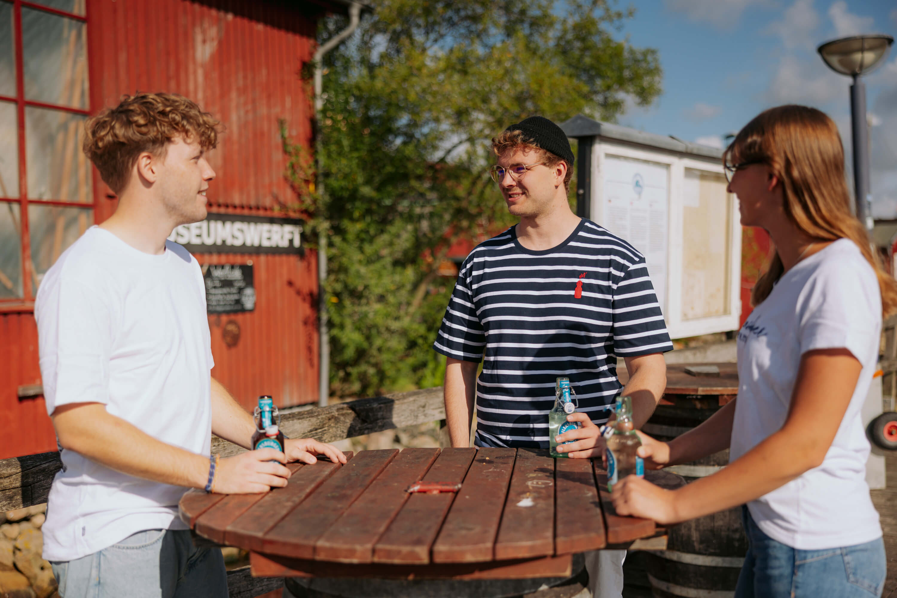 Drei Personen stehen an einem runden Holztisch im Freien und halten Flaschen mit blauer Etikette. Eine Person trägt ein blau-weiß gestreiftes Shirt mit rotem Motiv.