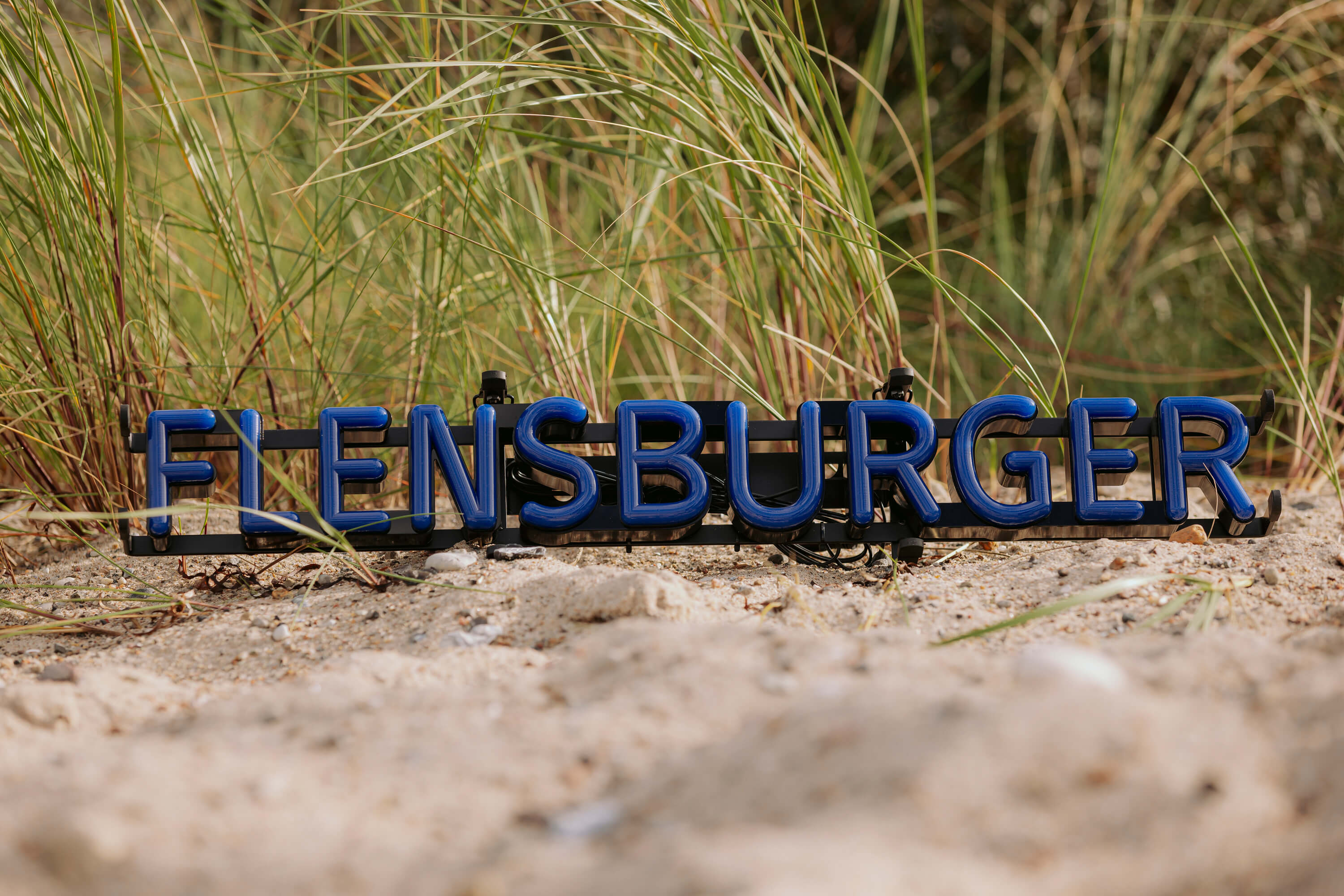 Das LED-Schild zeigt den Schriftzug "FLENSBURGER" in blauen Großbuchstaben auf schwarzem Rahmen. Es liegt auf Sand vor Hintergrund mit Gras.