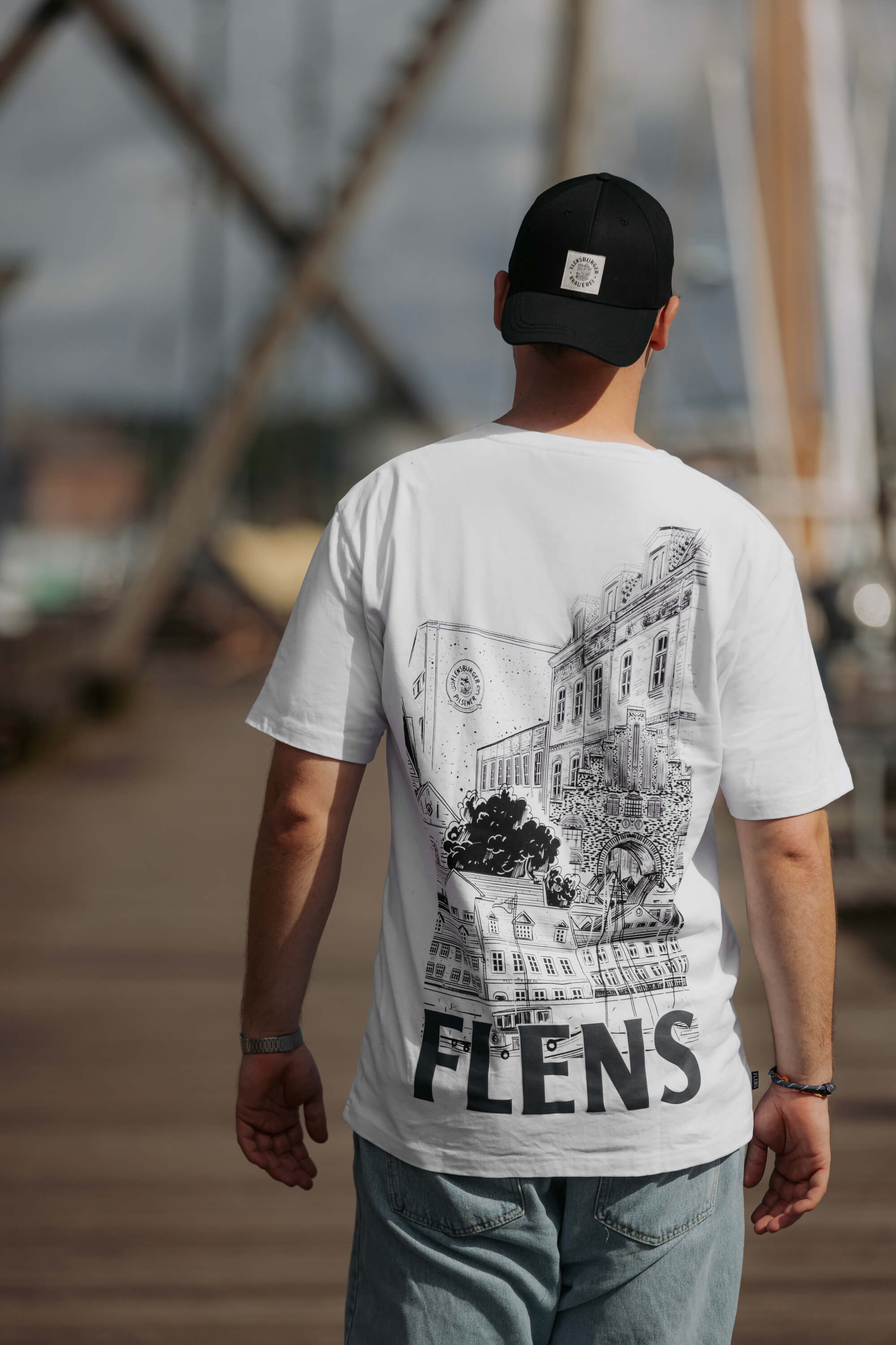 Mann trägt ein weißes Oversize-T-Shirt mit großem schwarzem Rückenprint von Flensburger Brauerei und dem Text "FLENS" in großen Buchstaben. Er trägt eine schwarze Kappe mit Logo.